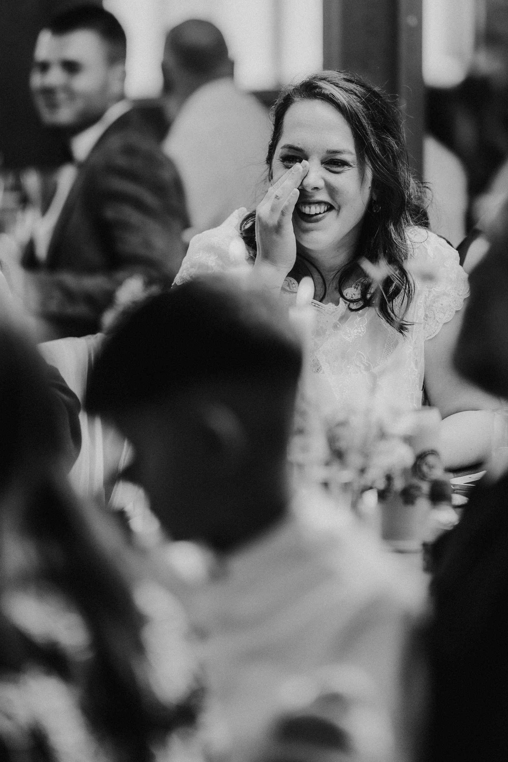 Daniel & Natalie’s Wedding at The Biscuit Factory 62 Bride laughing and wiping her eye at a wedding reception with guests blurred around her.
