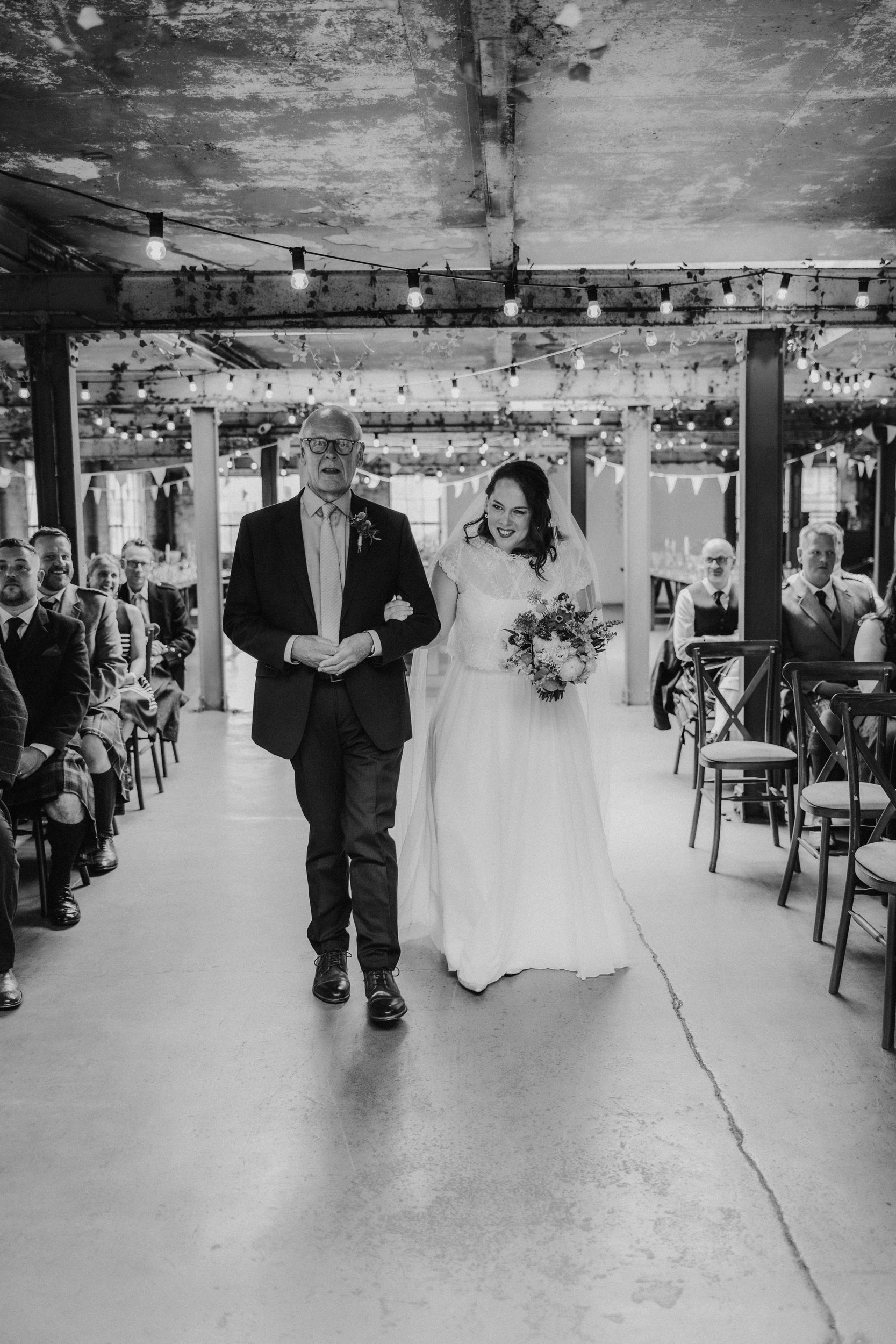 Daniel & Natalie’s Wedding at The Biscuit Factory 13 Bride Natalie arm-in-arm with an escort walking down an industrial aisle past seated guests and string lights.