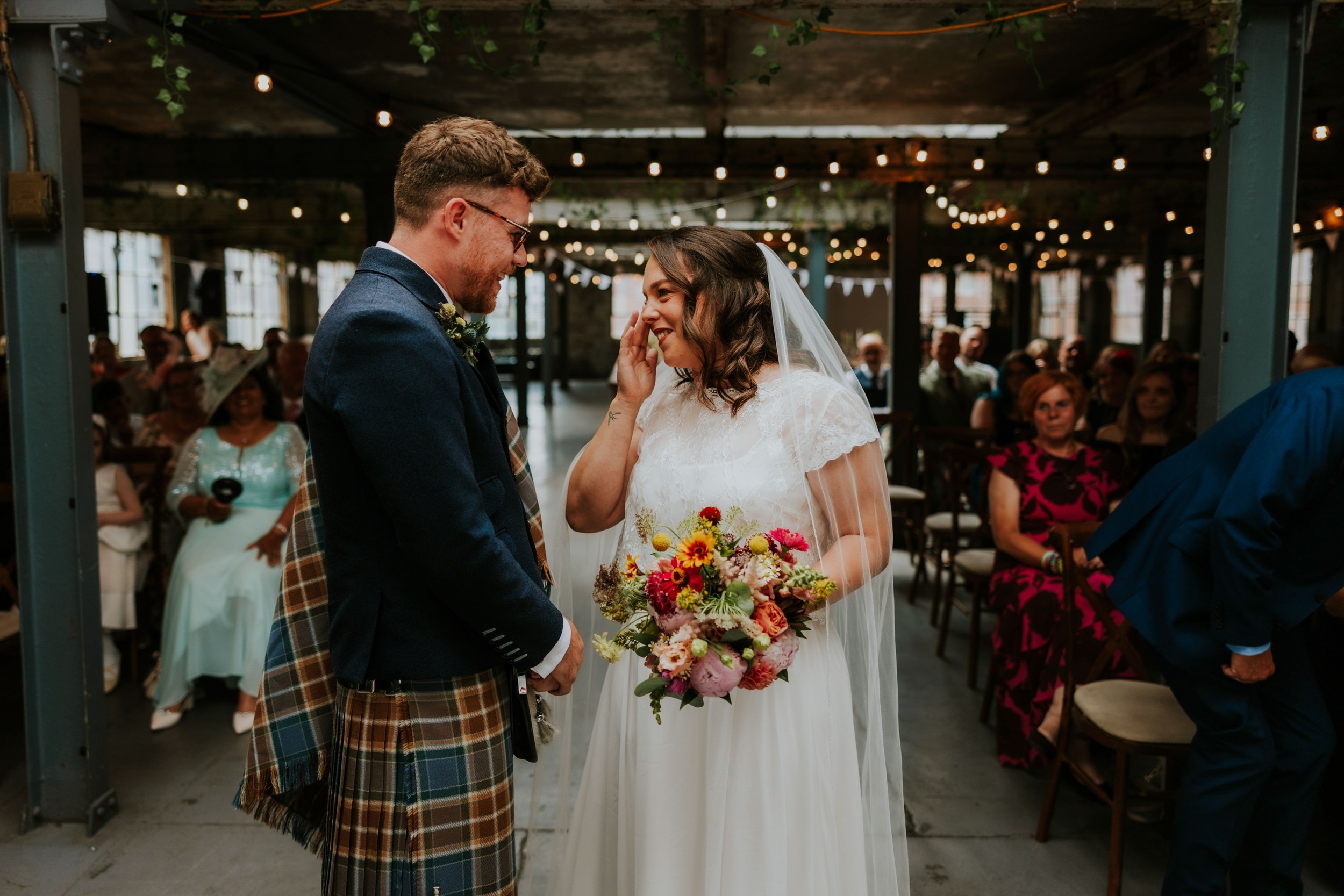 Daniel & Natalie’s Wedding at The Biscuit Factory 15 Bride wipes a tear facing groom in a kilt during a ceremony at The Biscuit Factory, holding a colourful bouquet under string lights.