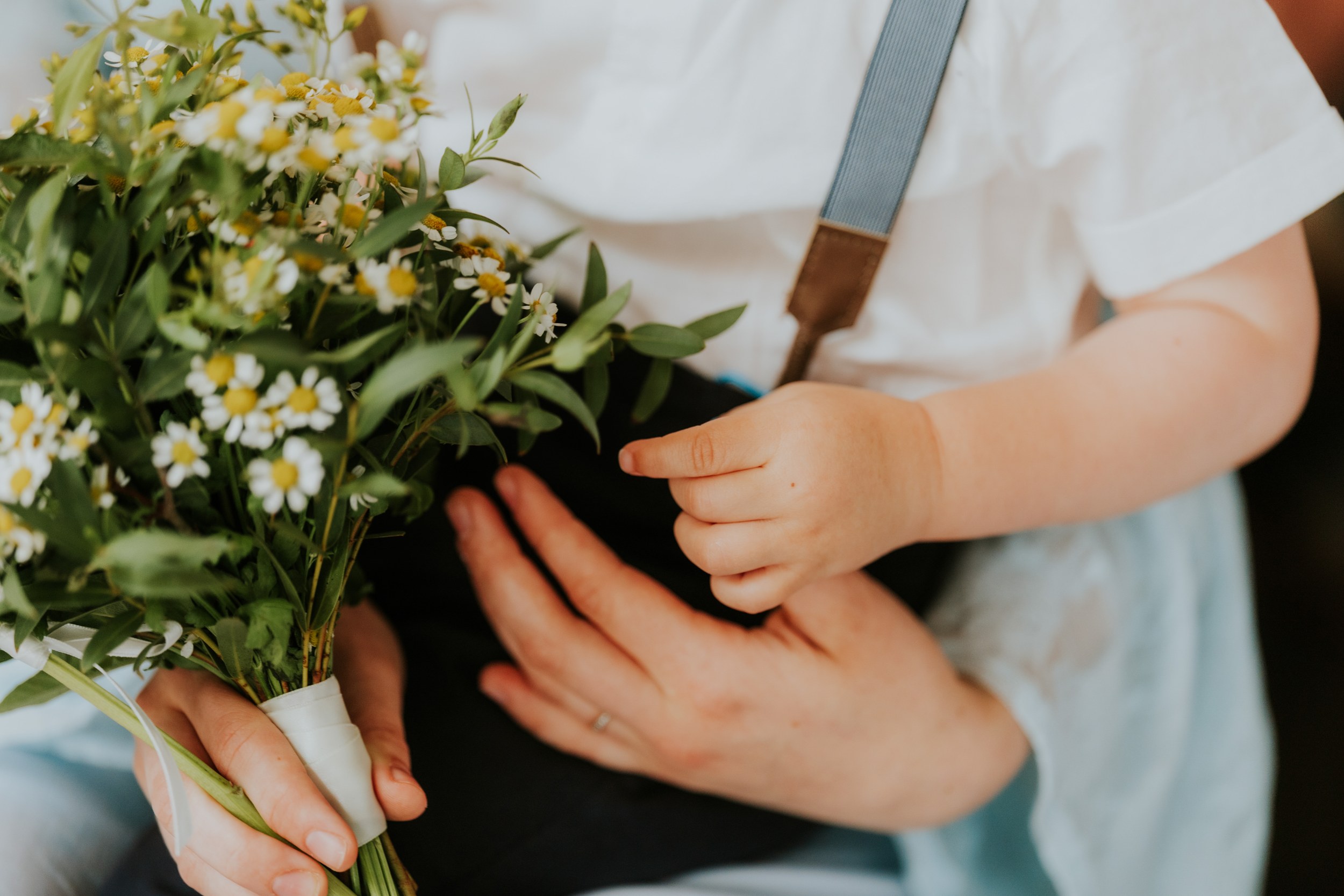 Daniel & Natalie’s Wedding at The Biscuit Factory 17 Close-up of adult and child hands holding a small daisy bouquet, child wearing a white top and grey-blue suspenders.
