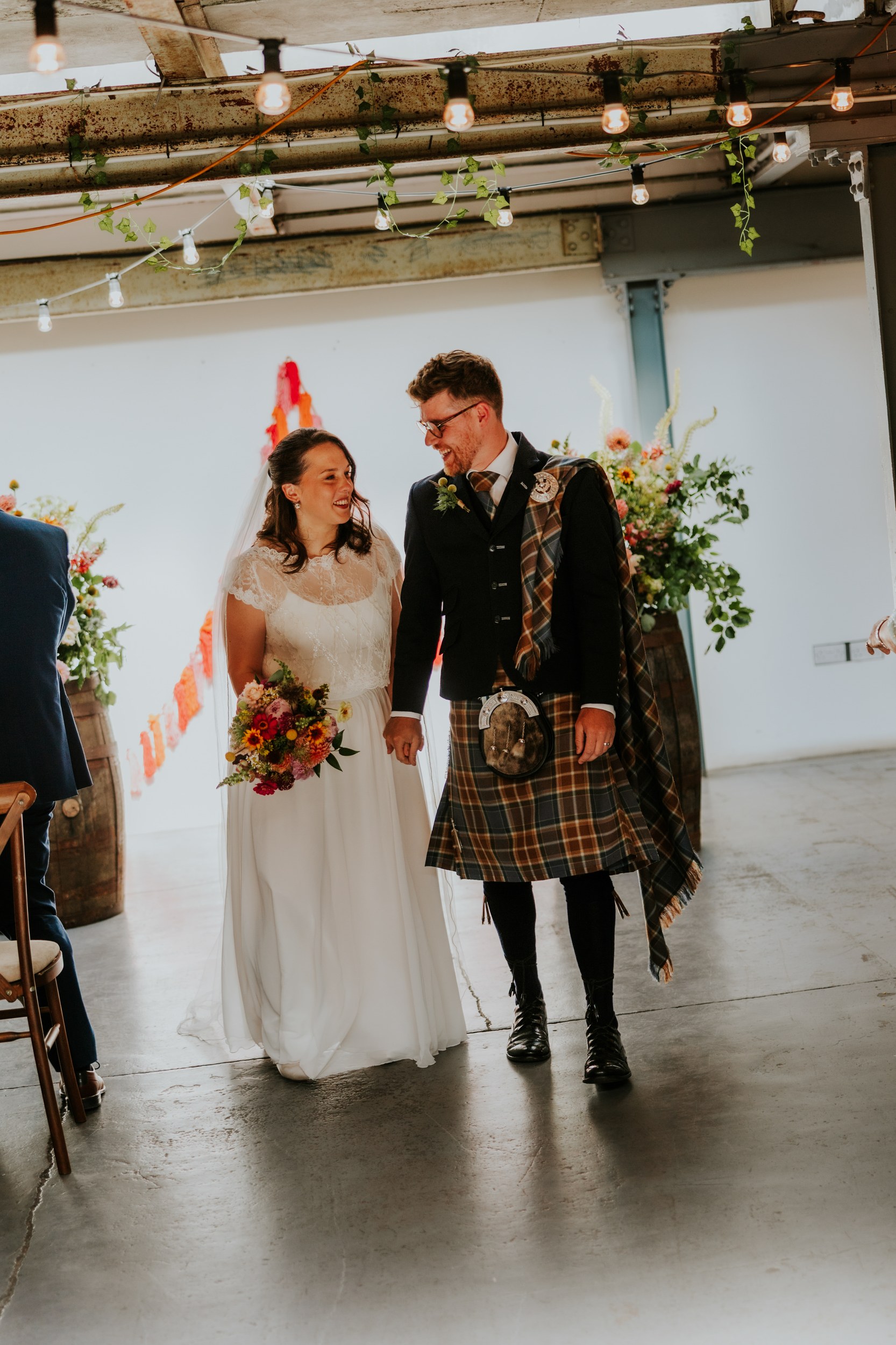Daniel & Natalie’s Wedding at The Biscuit Factory 30 Daniel and Natalie walking inside The Biscuit Factory, groom in tartan kilt and bride in lace dress holding a colourful bouquet under hanging bulbs.