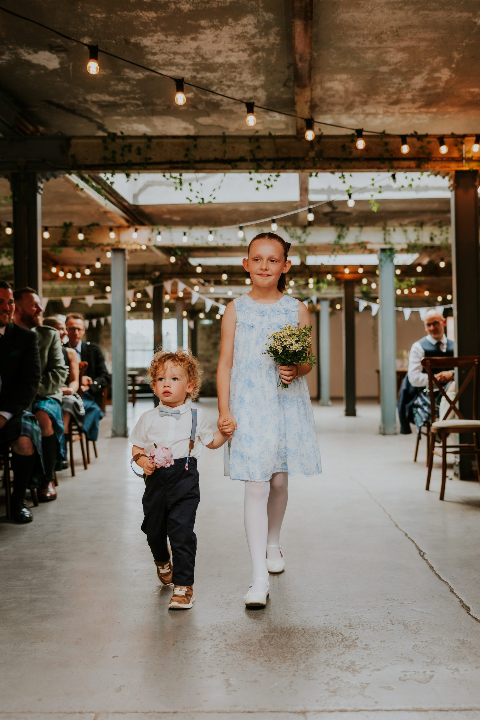 Daniel & Natalie’s Wedding at The Biscuit Factory 12 Flower girl in a pale blue dress with bouquet and page boy in suspenders walk hand‑in‑hand down a string‑lit aisle at The Biscuit Factory.