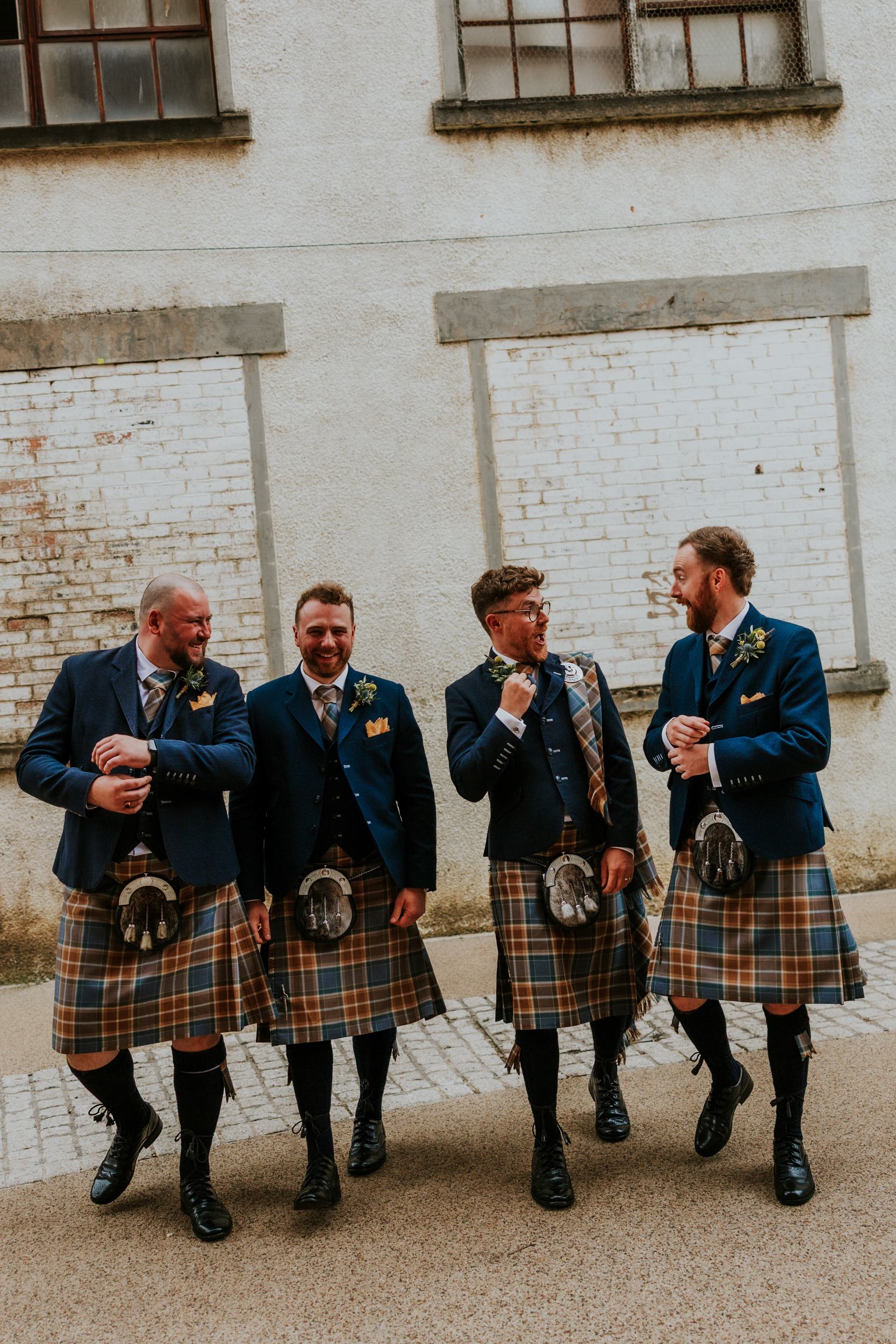 Daniel & Natalie’s Wedding at The Biscuit Factory 39 Four groomsmen in navy jackets and tartan kilts laughing outside a white brick wall.