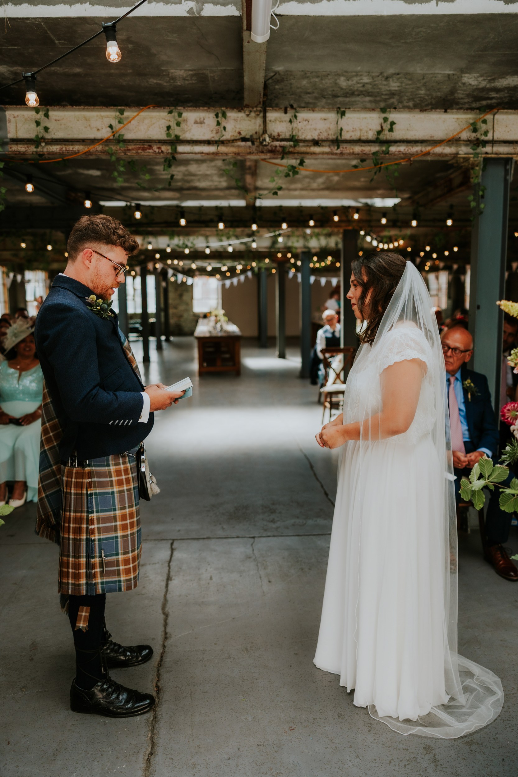 Daniel & Natalie’s Wedding at The Biscuit Factory 24 Groom in tartan kilt reads vows to bride in veil during indoor ceremony at The Biscuit Factory with string lights.