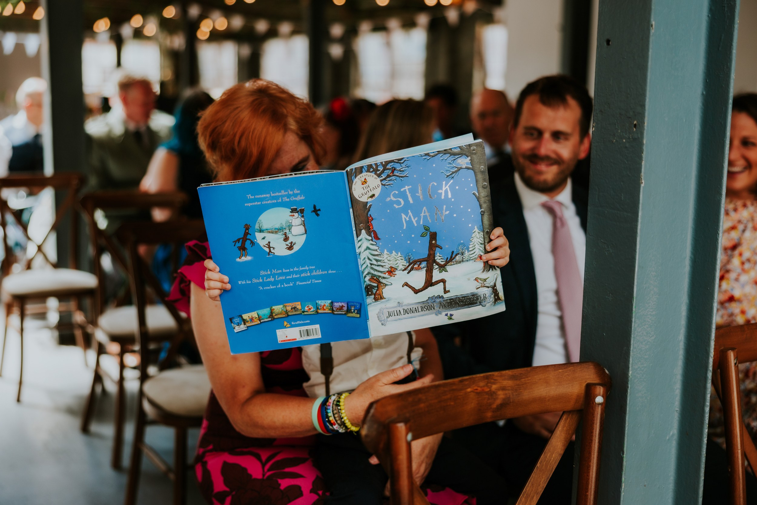 Daniel & Natalie’s Wedding at The Biscuit Factory 11 Guest hiding behind the children's book 'Stick Man' during a seated wedding ceremony