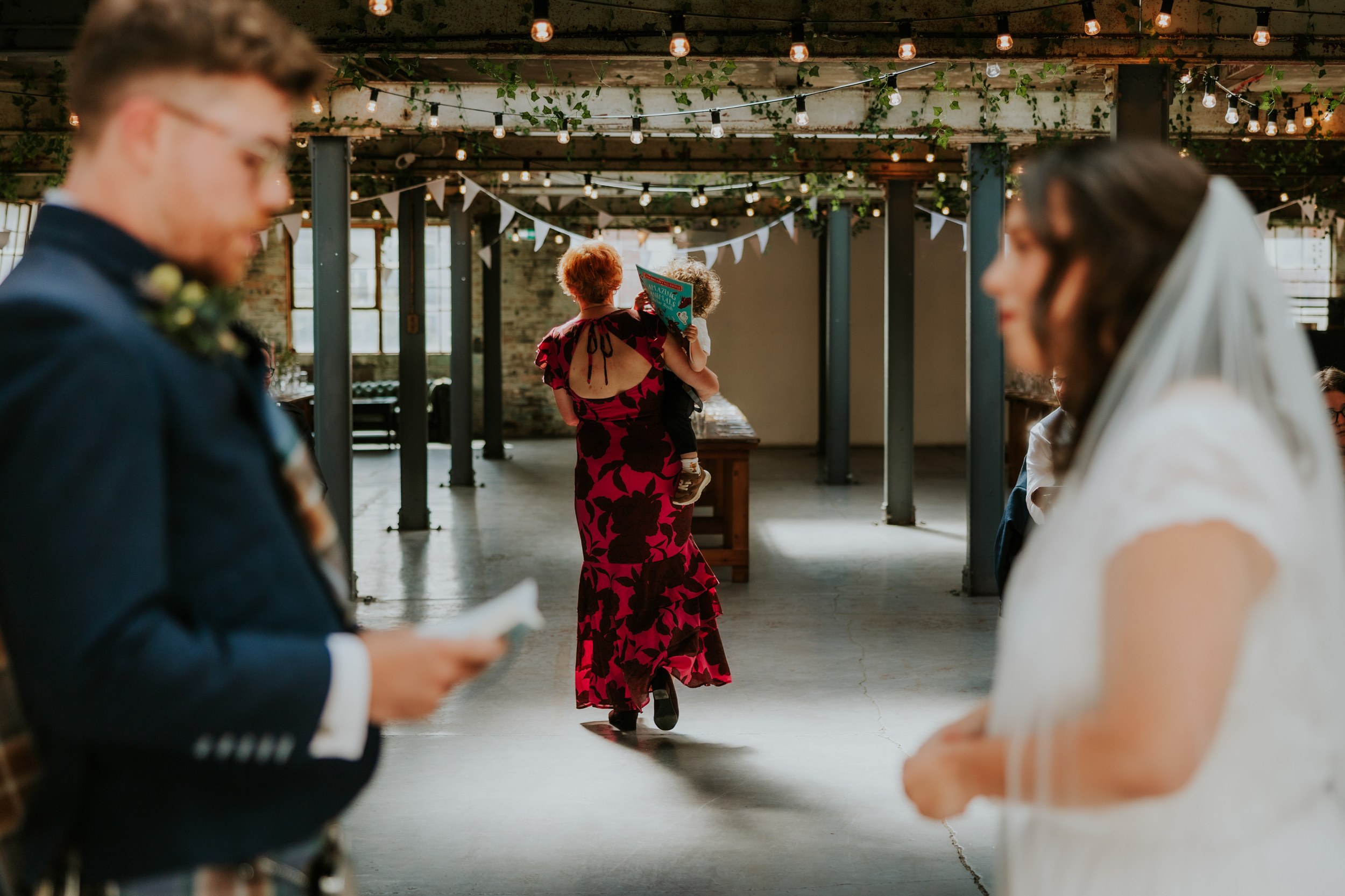 Daniel & Natalie’s Wedding at The Biscuit Factory 23 Guest in red dress carrying a child with a book walks through industrial venue decorated with string lights and bunting; bride and groom blurred.