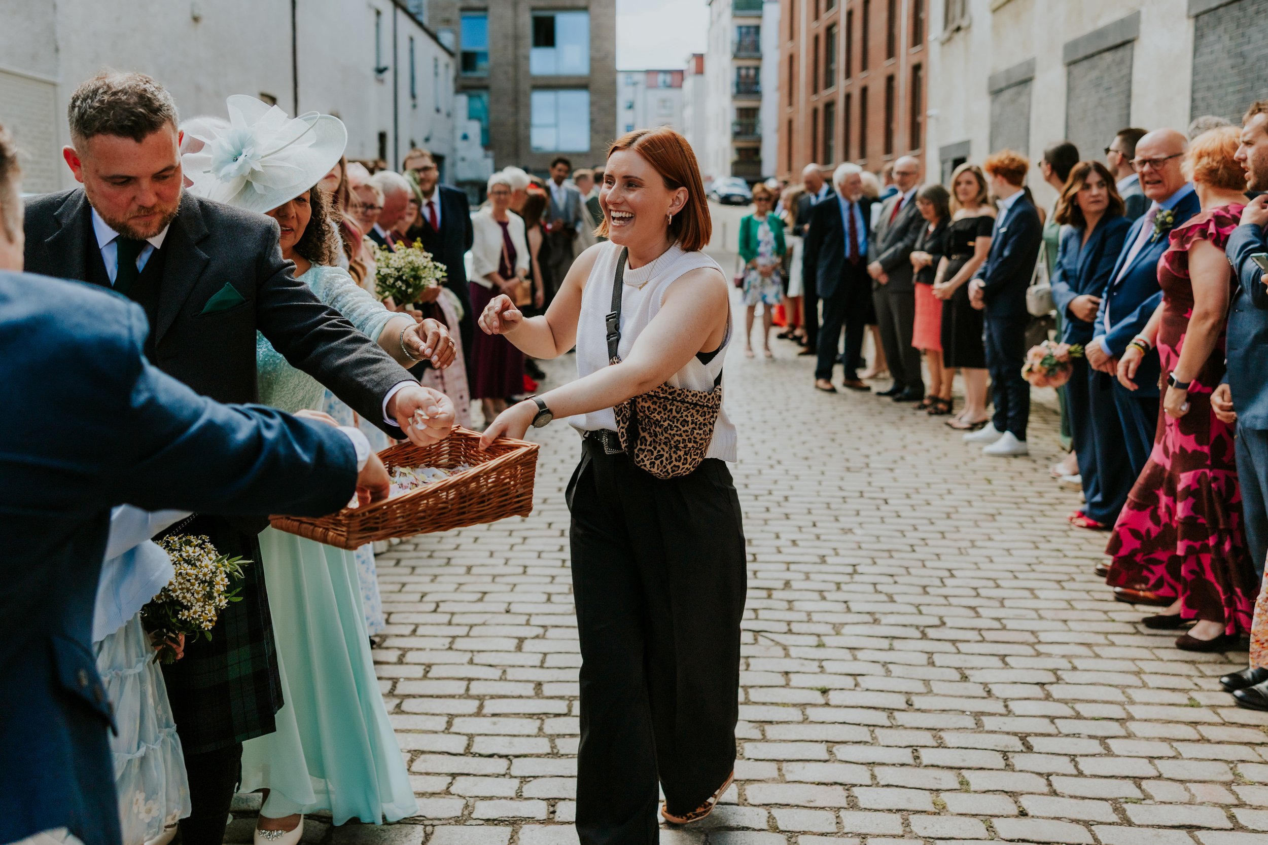 Daniel & Natalie’s Wedding at The Biscuit Factory 31 Guest smiling as she takes confetti from a wicker basket while wedding guests line a cobbled street outside The Biscuit Factory, Edinburgh.