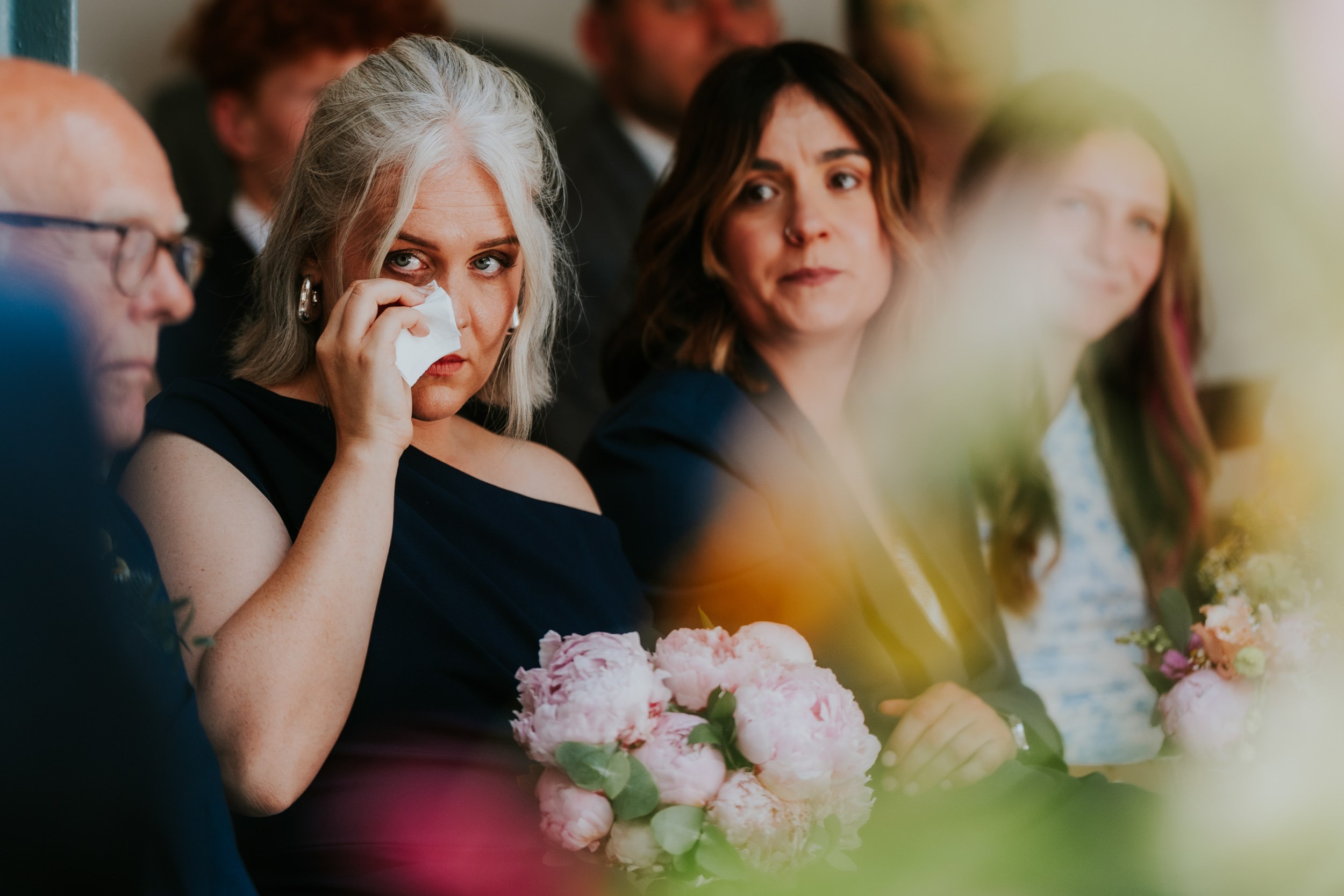 Daniel & Natalie’s Wedding at The Biscuit Factory 19 Guest wiping tears with a tissue at a wedding, holding a pink peony bouquet, other guests blurred.