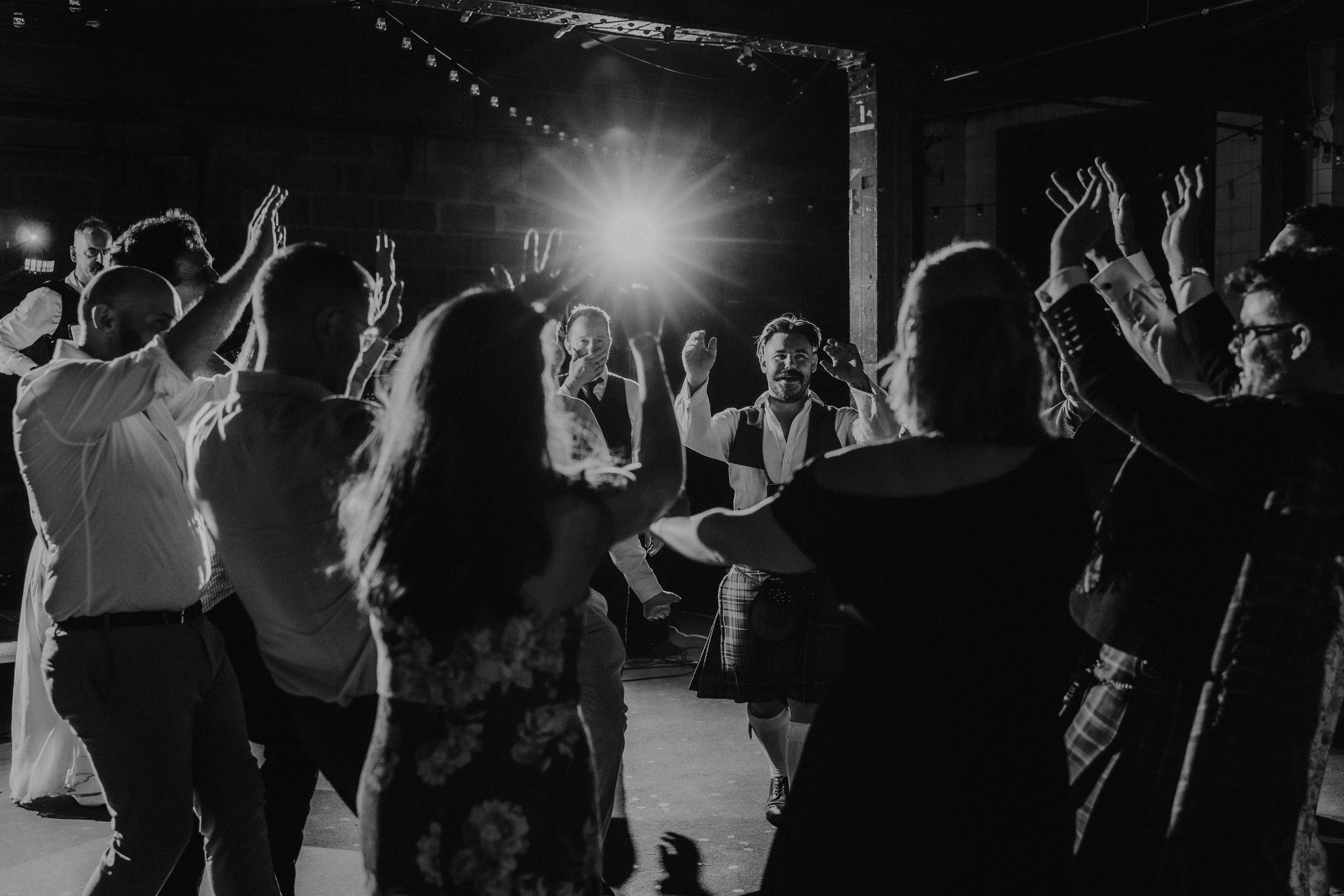 Daniel & Natalie’s Wedding at The Biscuit Factory 79 Guests dancing on a dark reception dancefloor, backlit by a bright flash; a man in a kilt raises his hands.