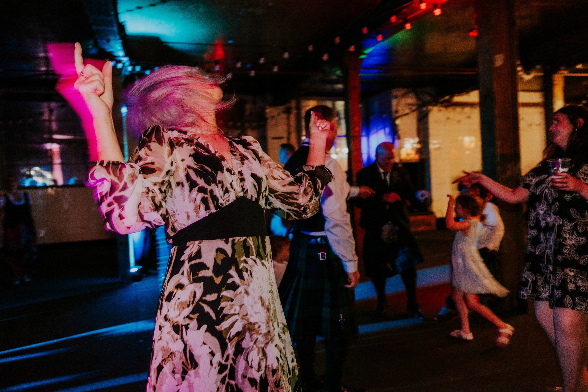Daniel & Natalie’s Wedding at The Biscuit Factory 87 Guests dancing under coloured lights at a wedding reception; woman in patterned dress with arms raised.
