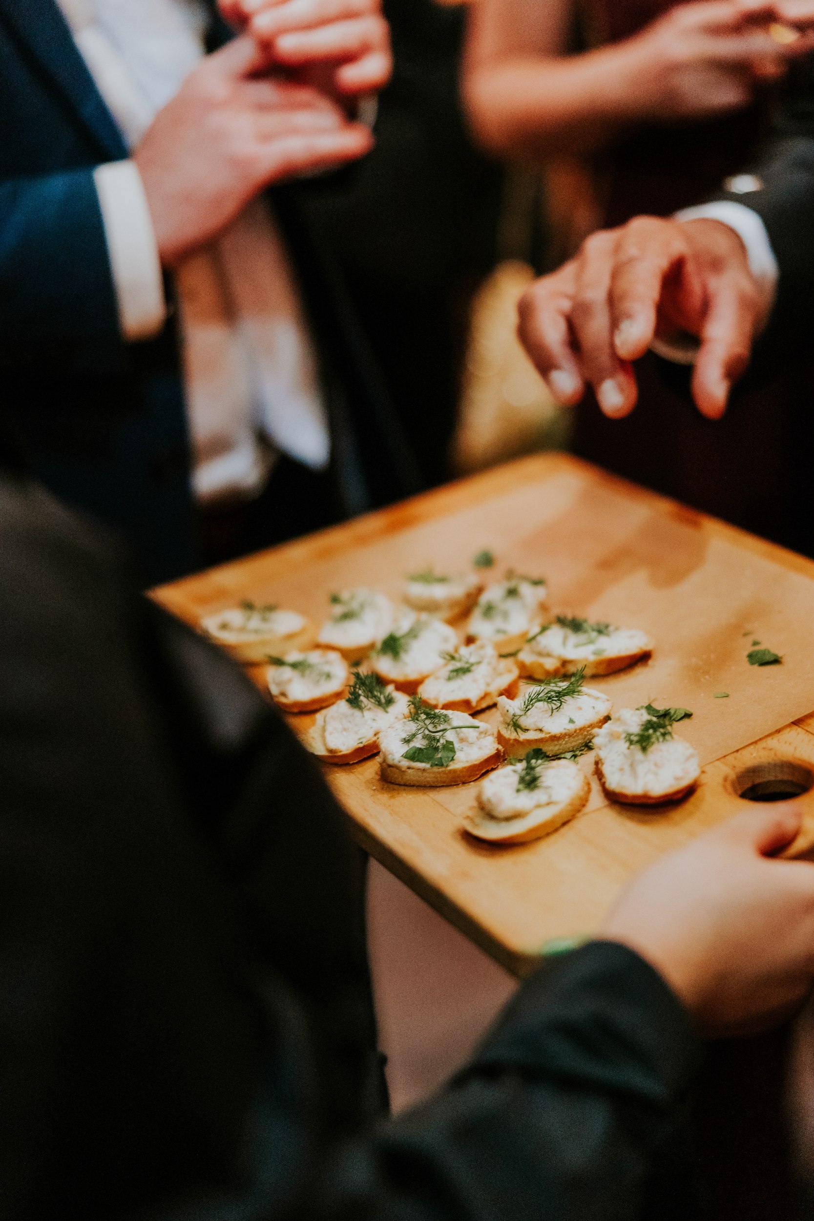 Daniel & Natalie’s Wedding at The Biscuit Factory 36 Guests in suits reaching for canapés on a wooden serving board at a wedding reception.