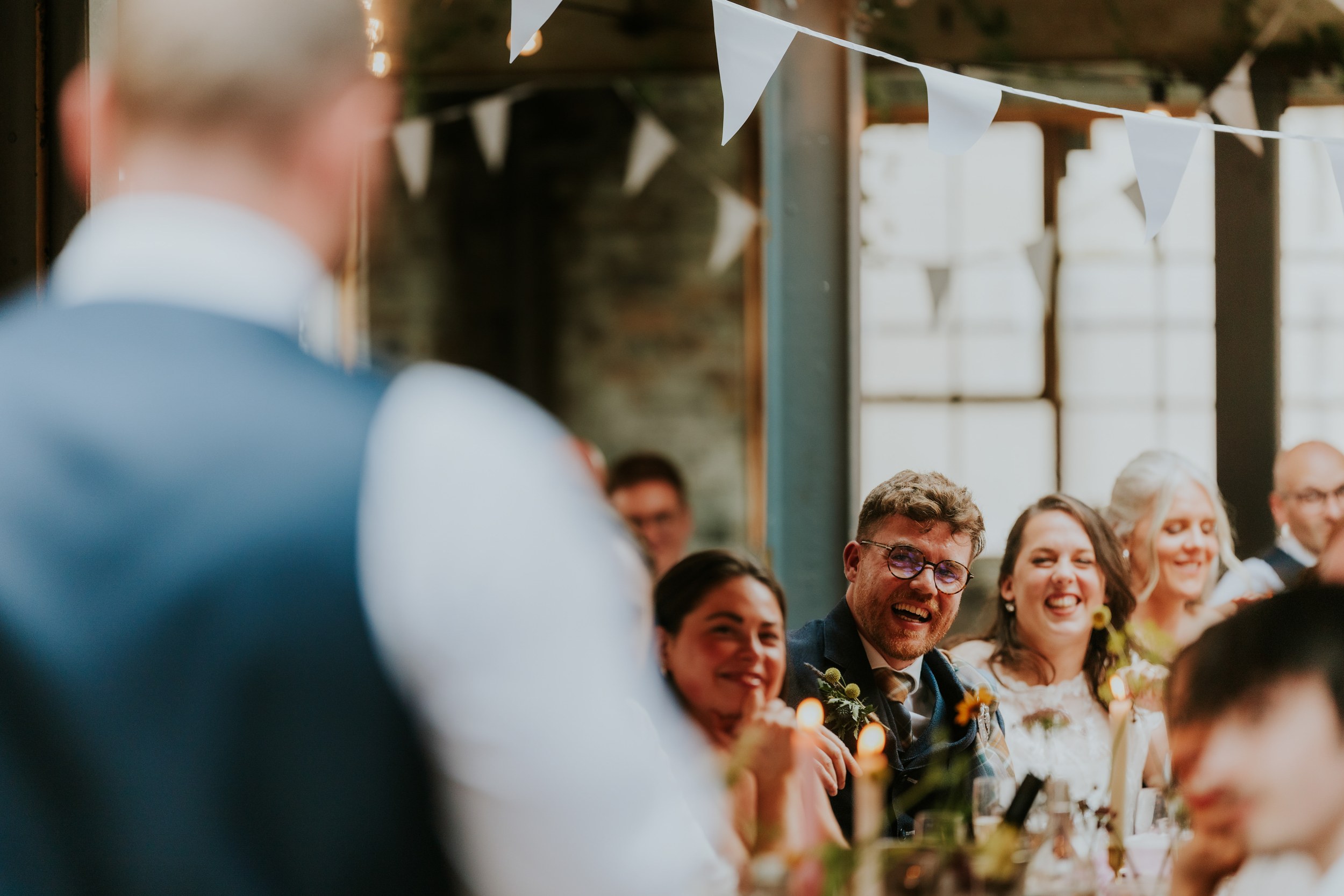 Daniel & Natalie’s Wedding at The Biscuit Factory 65 Guests smiling and laughing at a wedding speech with bunting and candles in an industrial-style room.