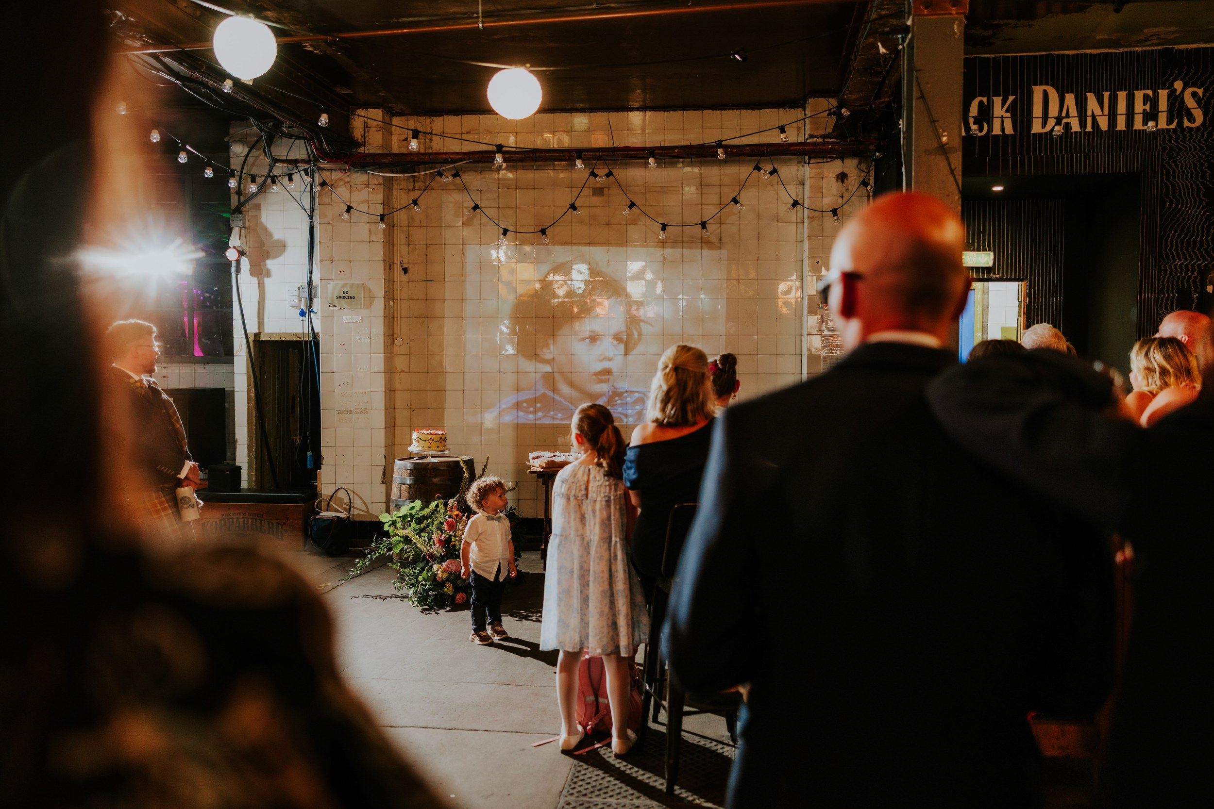 Daniel & Natalie’s Wedding at The Biscuit Factory 68 Guests watch a projected image on a tiled wall at The Biscuit Factory; cake on a barrel and children stand in the foreground.