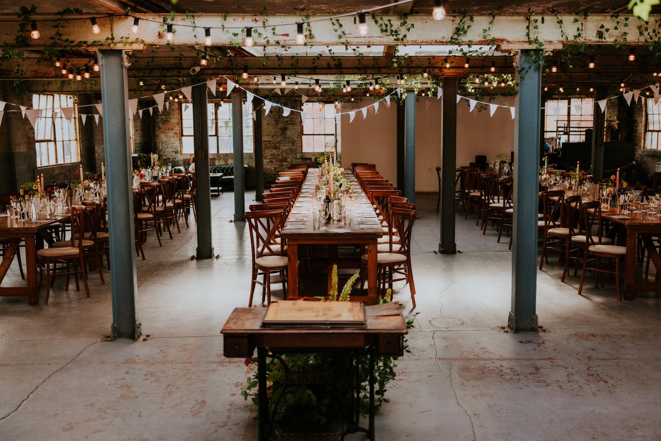 Daniel & Natalie’s Wedding at The Biscuit Factory 53 Long banquet tables and wooden chairs set for a wedding reception in an industrial loft with bunting, string lights and exposed brick windows.