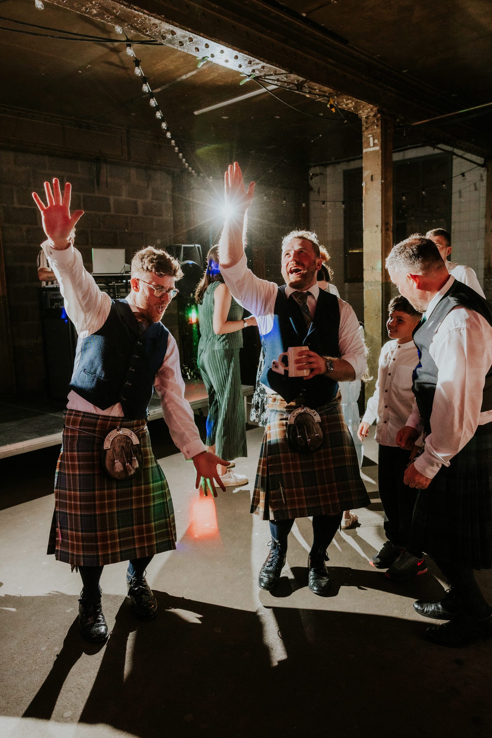 Daniel & Natalie’s Wedding at The Biscuit Factory 85 Male guests in kilts dancing under string lights at a reception in The Biscuit Factory, Edinburgh.