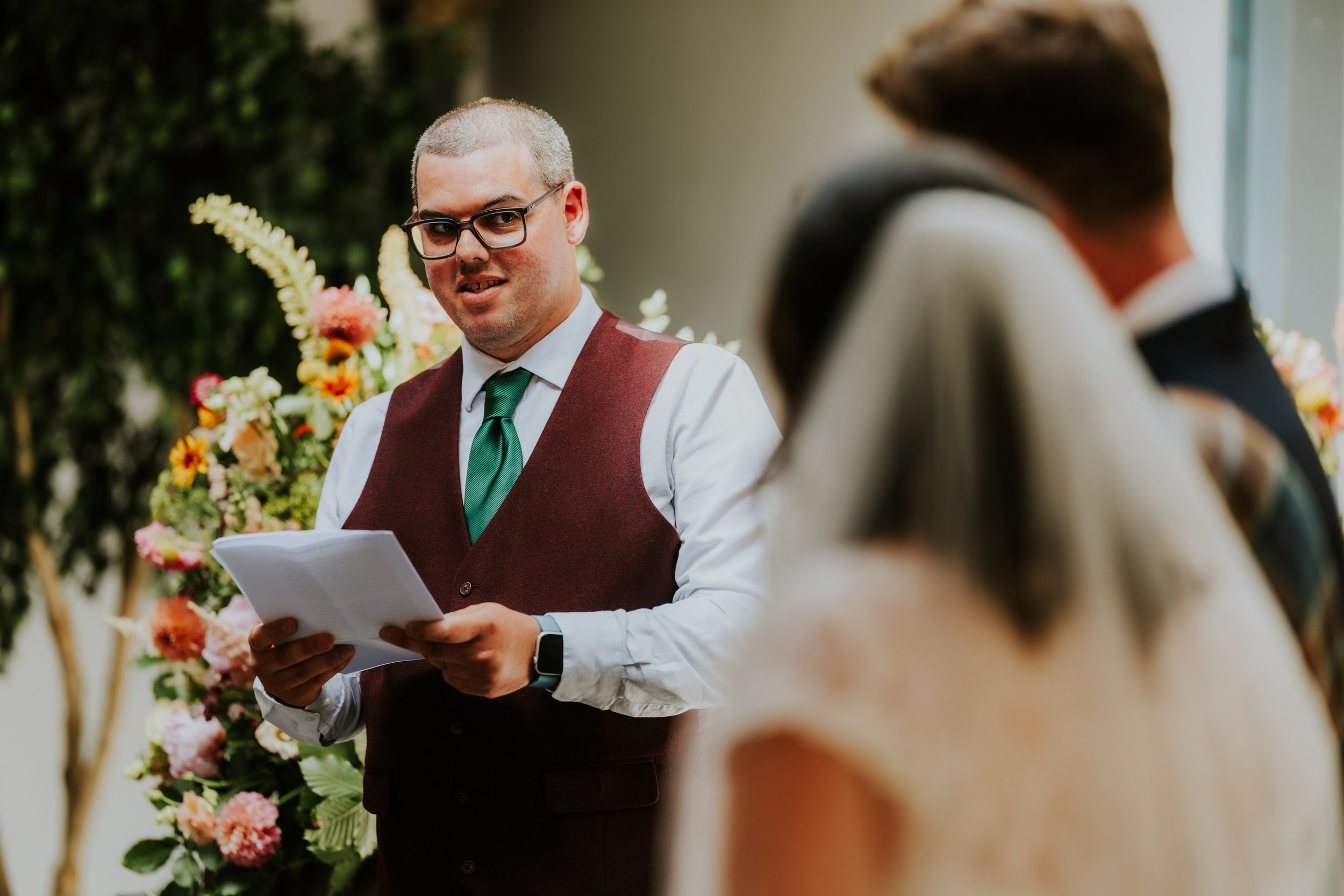 Daniel & Natalie’s Wedding at The Biscuit Factory 20 Man in glasses and burgundy waistcoat reading from papers at an indoor wedding with floral backdrop.