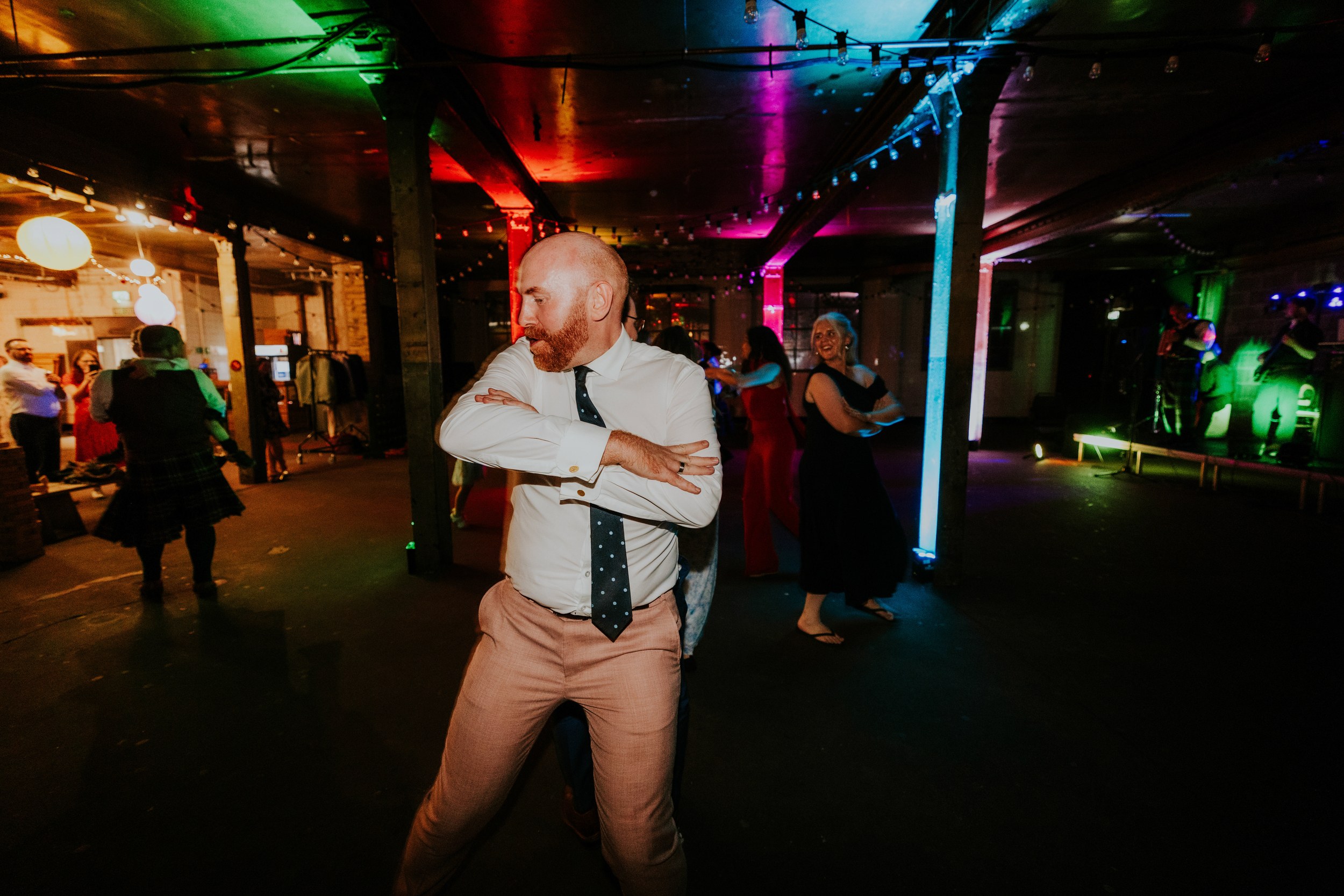 Daniel & Natalie’s Wedding at The Biscuit Factory 78 Man in shirt and tie dancing on a dim, colourfully lit dance floor at The Biscuit Factory with guests and a live band.