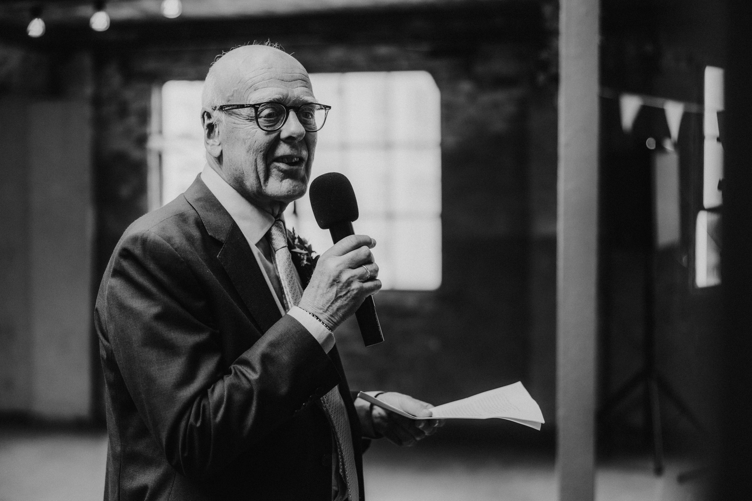 Daniel & Natalie’s Wedding at The Biscuit Factory 61 Man in suit and glasses holding a microphone and notes, speaking inside The Biscuit Factory.