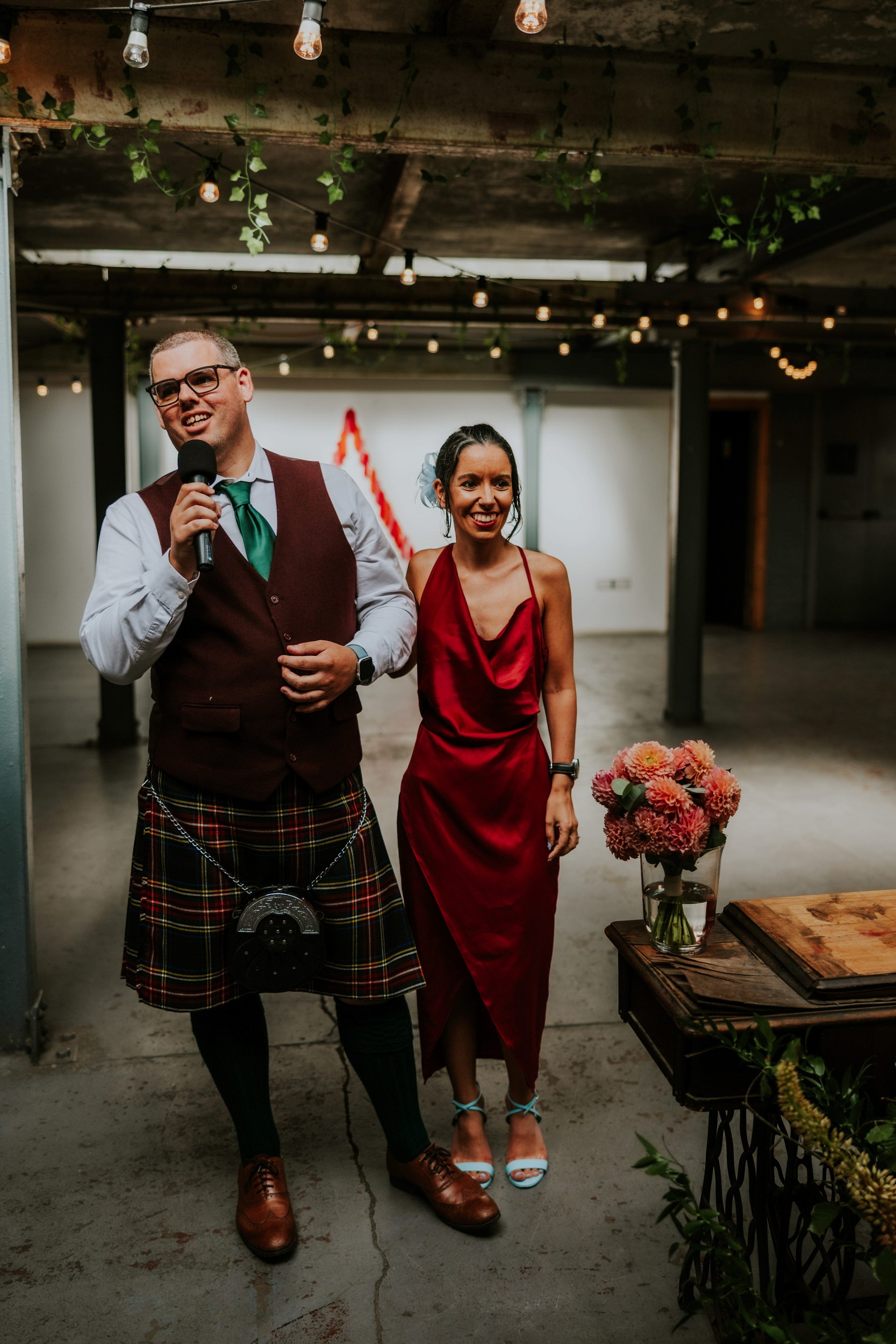 Daniel & Natalie’s Wedding at The Biscuit Factory 59 Man in tartan kilt holding a microphone with a woman in a red dress smiling beside him under string lights and a vase of flowers.