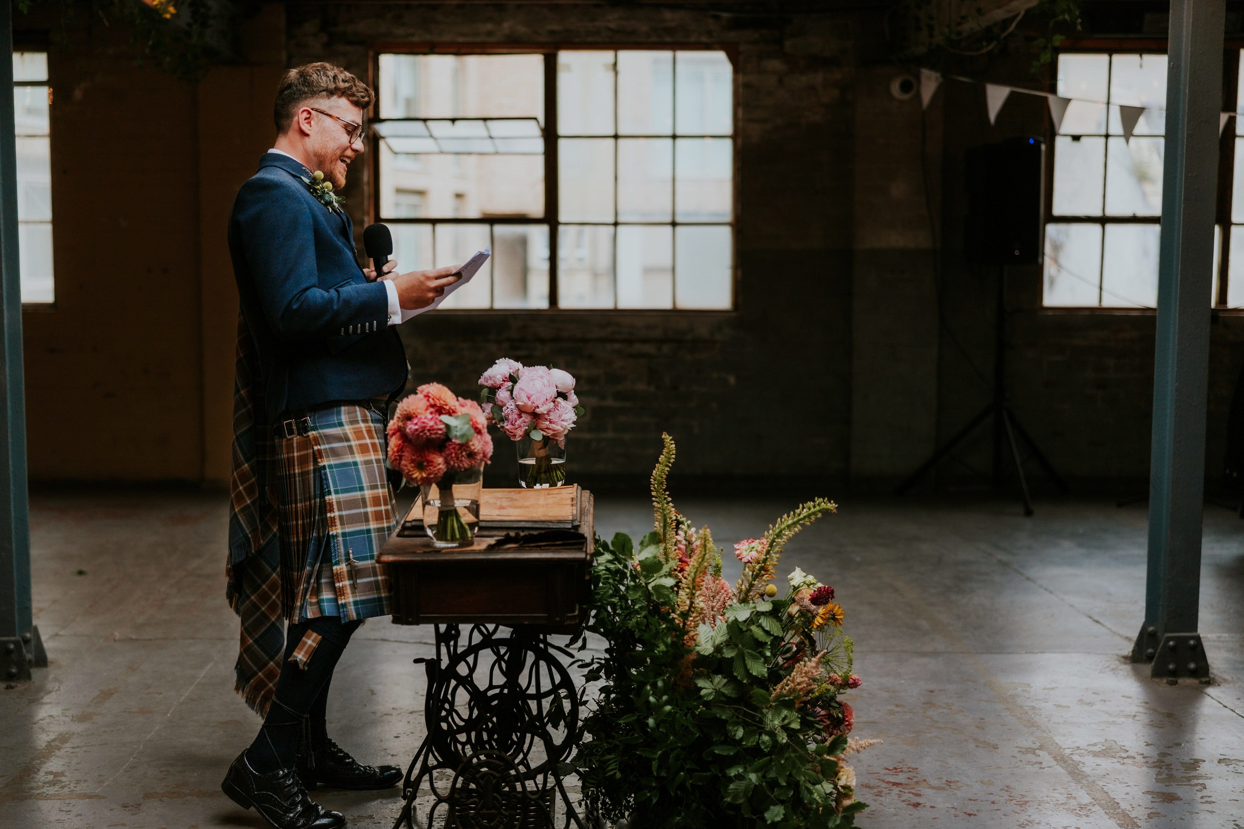 Daniel & Natalie’s Wedding at The Biscuit Factory 66 Man in tartan kilt reads a speech into a microphone beside floral arrangements in an industrial wedding venue.