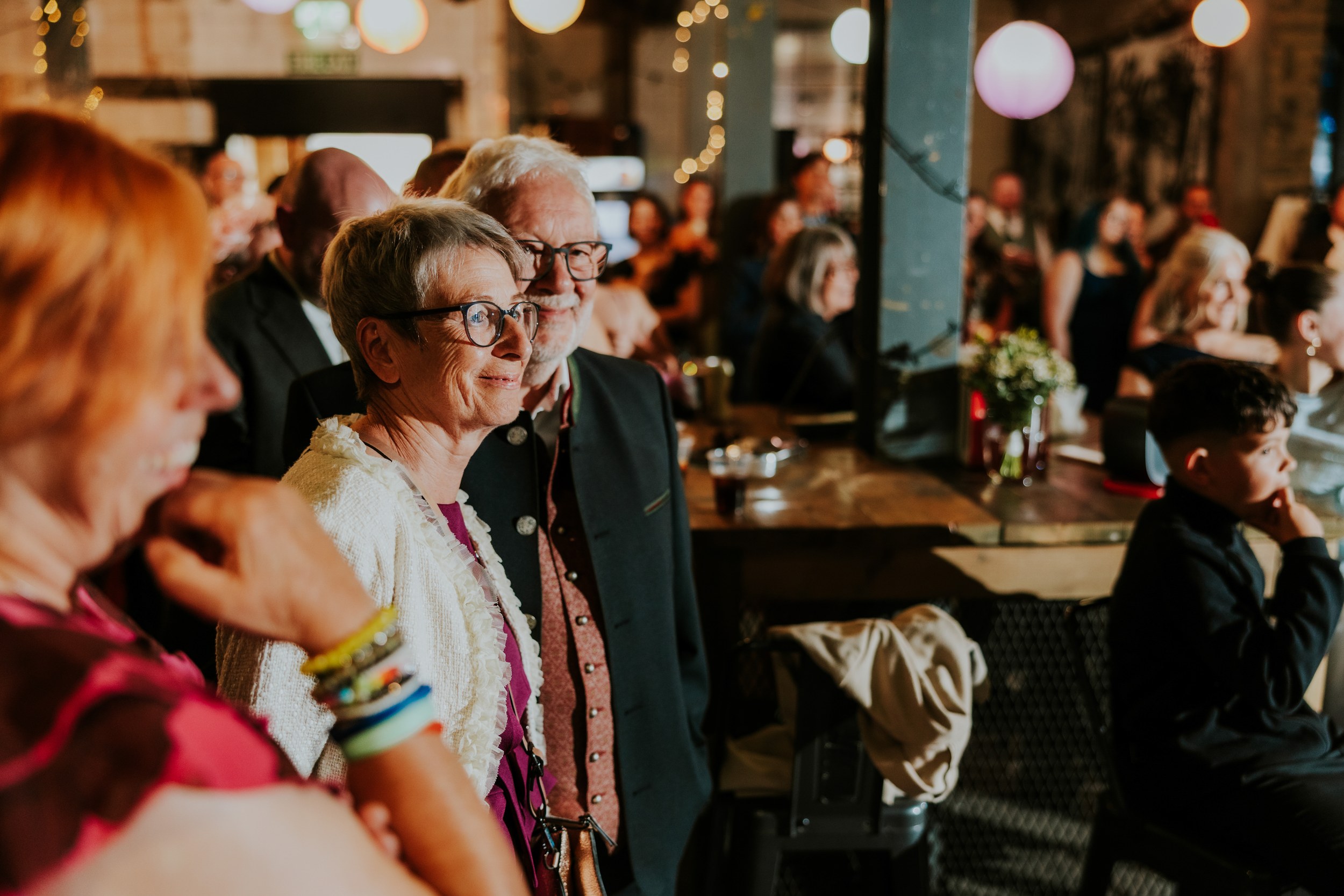 Daniel & Natalie’s Wedding at The Biscuit Factory 69 Older couple smiling among wedding guests in an industrial-style reception space with warm lights.