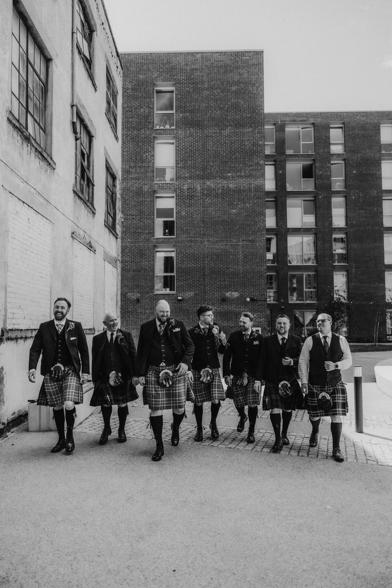 Daniel & Natalie’s Wedding at The Biscuit Factory 3 Seven groomsmen in kilts walking and laughing outside a brick industrial building at The Biscuit Factory, Edinburgh.