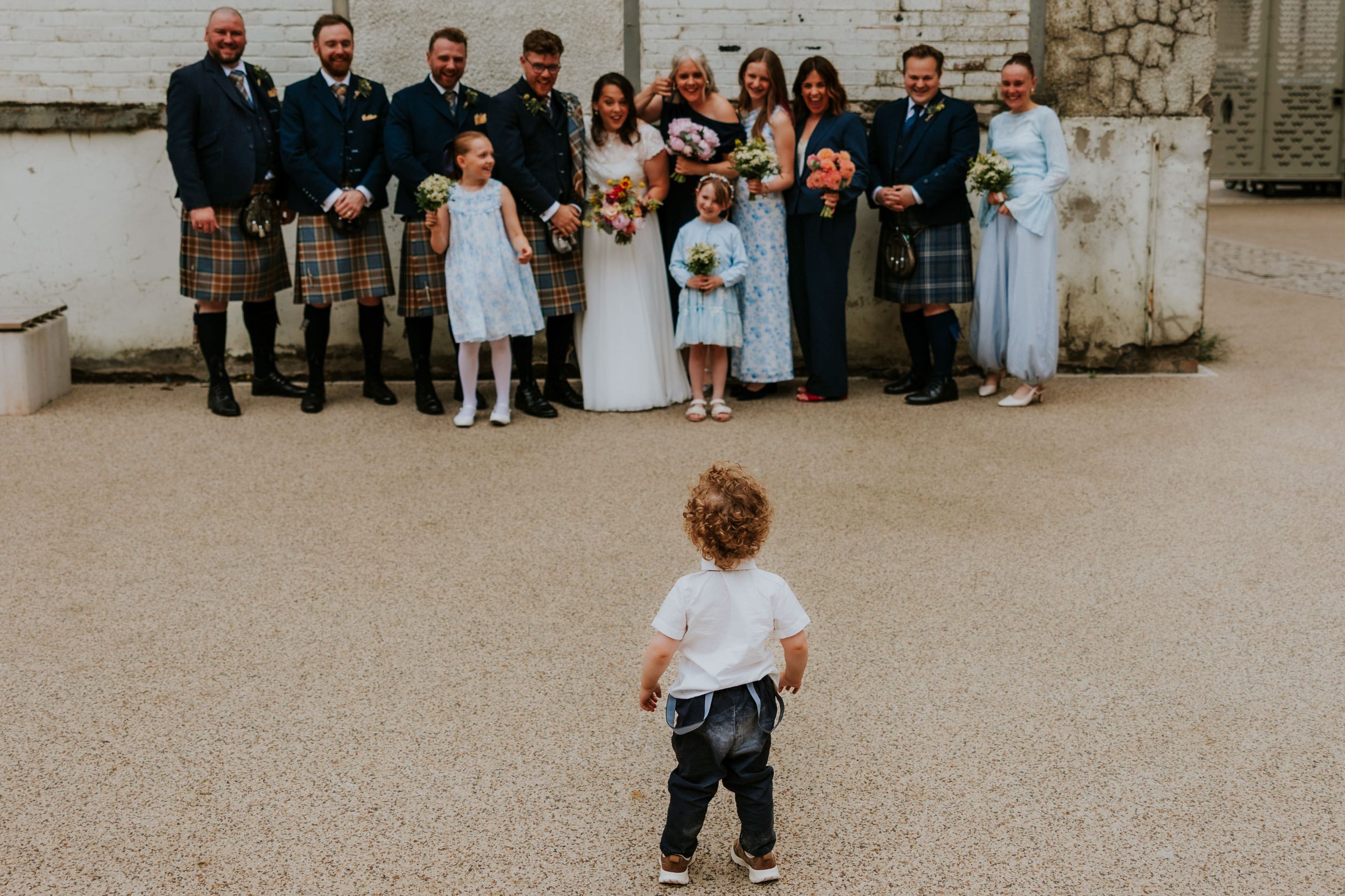 Daniel & Natalie’s Wedding at The Biscuit Factory 40 Small boy approaches a wedding party by a white brick wall; bride in white and groomsmen in kilts.