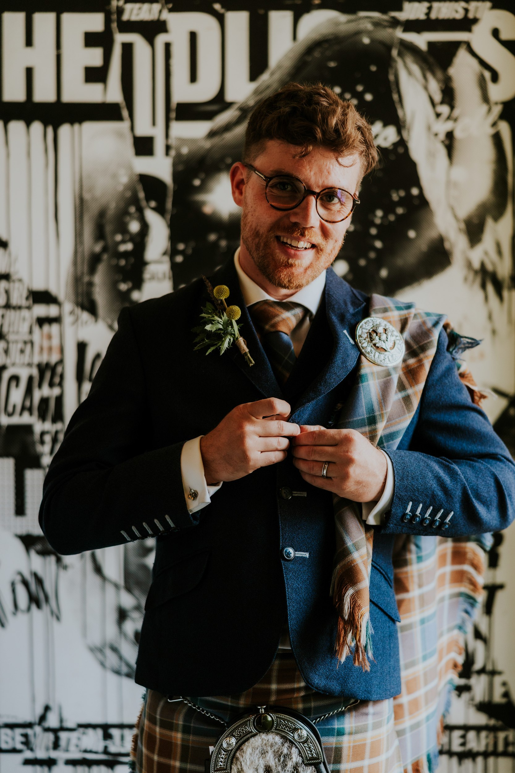 Daniel & Natalie’s Wedding at The Biscuit Factory 51 Smiling groom in glasses wearing kilt, tartan sash and sporran, adjusting his jacket in front of a black-and-white mural.