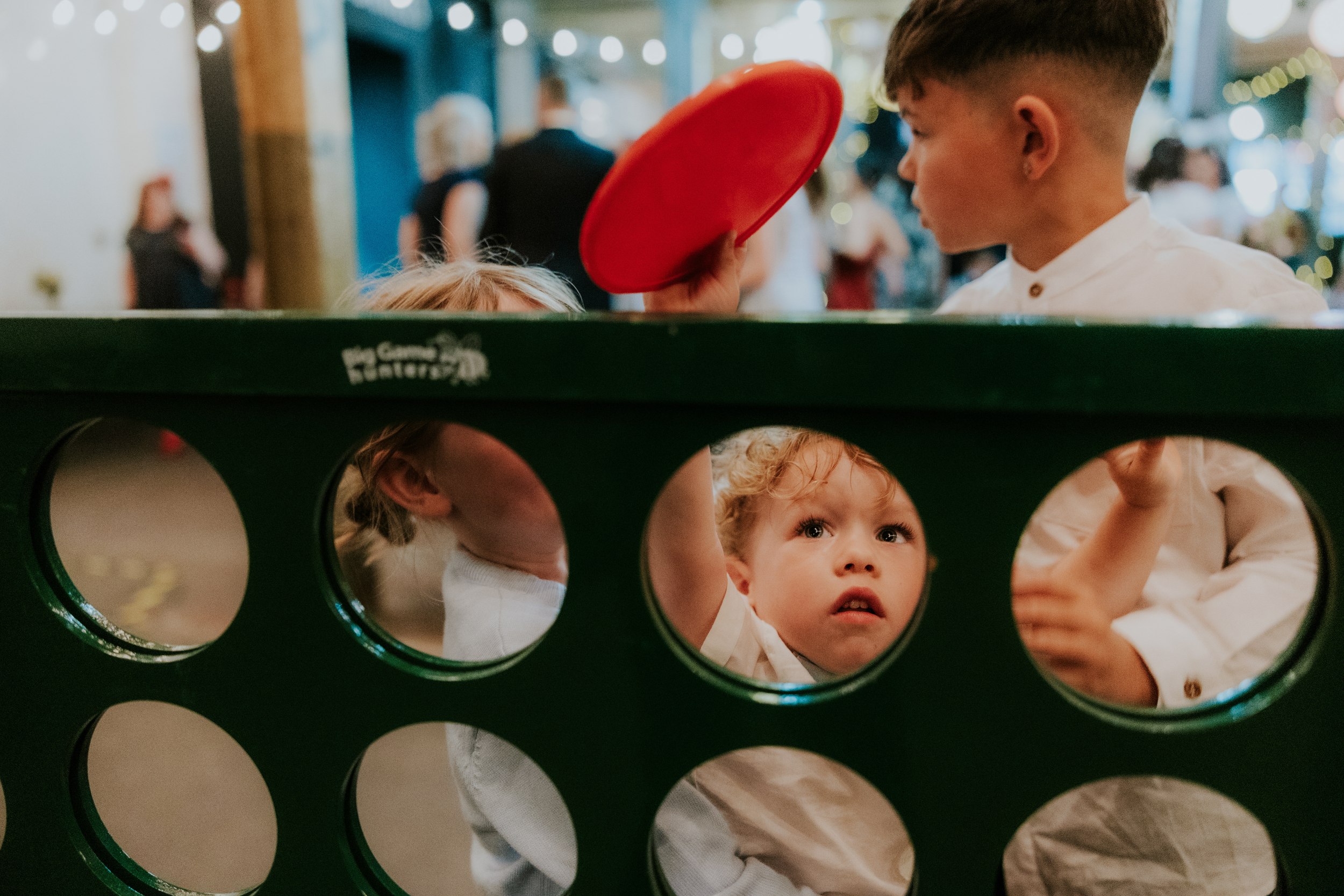 Daniel & Natalie’s Wedding at The Biscuit Factory 35 Three children peering through a green game board as a boy reaches up with a red disc, blurred guests and lights behind.