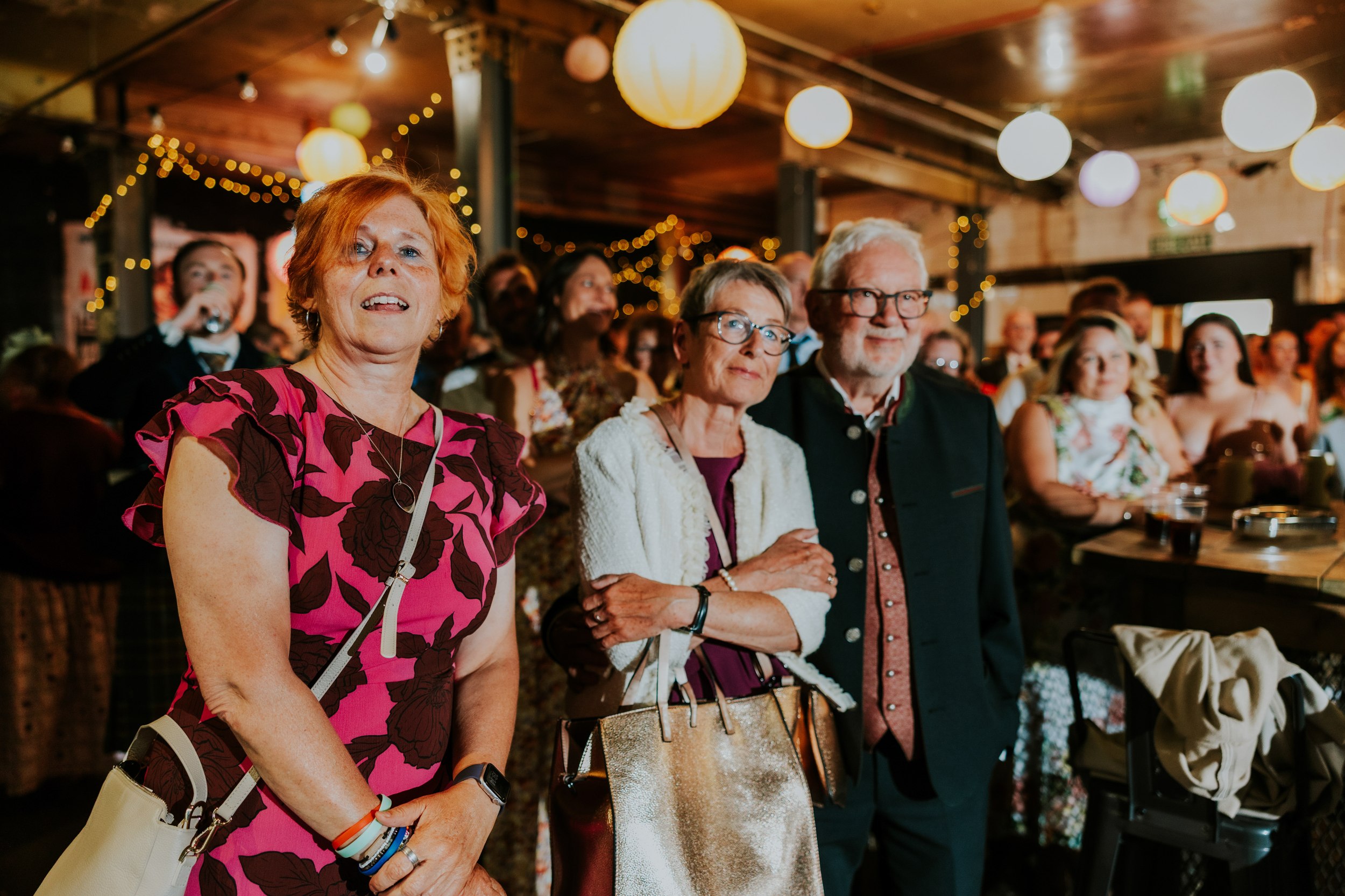 Daniel & Natalie’s Wedding at The Biscuit Factory 70 Three wedding guests watching a speech indoors under string lights and paper lanterns.
