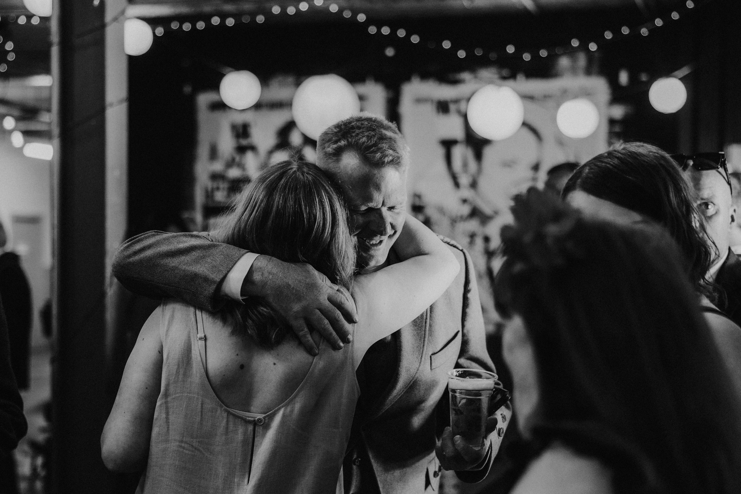 Daniel & Natalie’s Wedding at The Biscuit Factory 4 Two guests embrace at a wedding reception under hanging string lights, a man holding a pint.