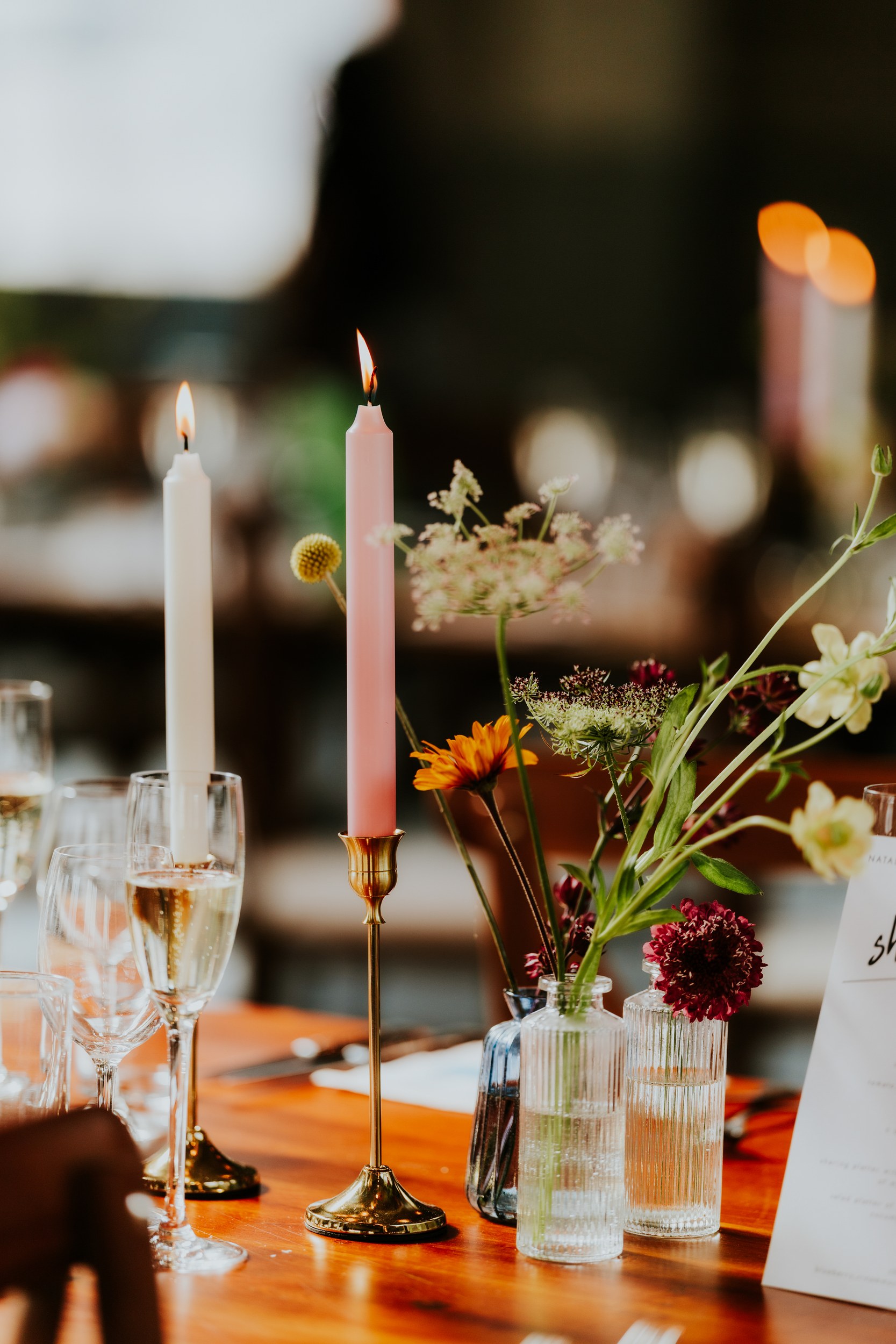 Daniel & Natalie’s Wedding at The Biscuit Factory 55 Two lit taper candles and small vases of flowers on a wooden wedding reception table.
