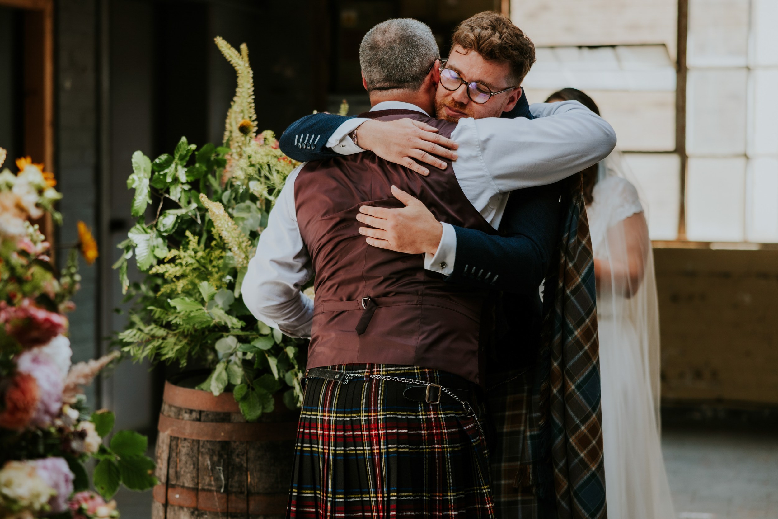 Daniel & Natalie’s Wedding at The Biscuit Factory 22 Two men in formal wear embrace at an indoor wedding, with flowers and a veiled bride visible.