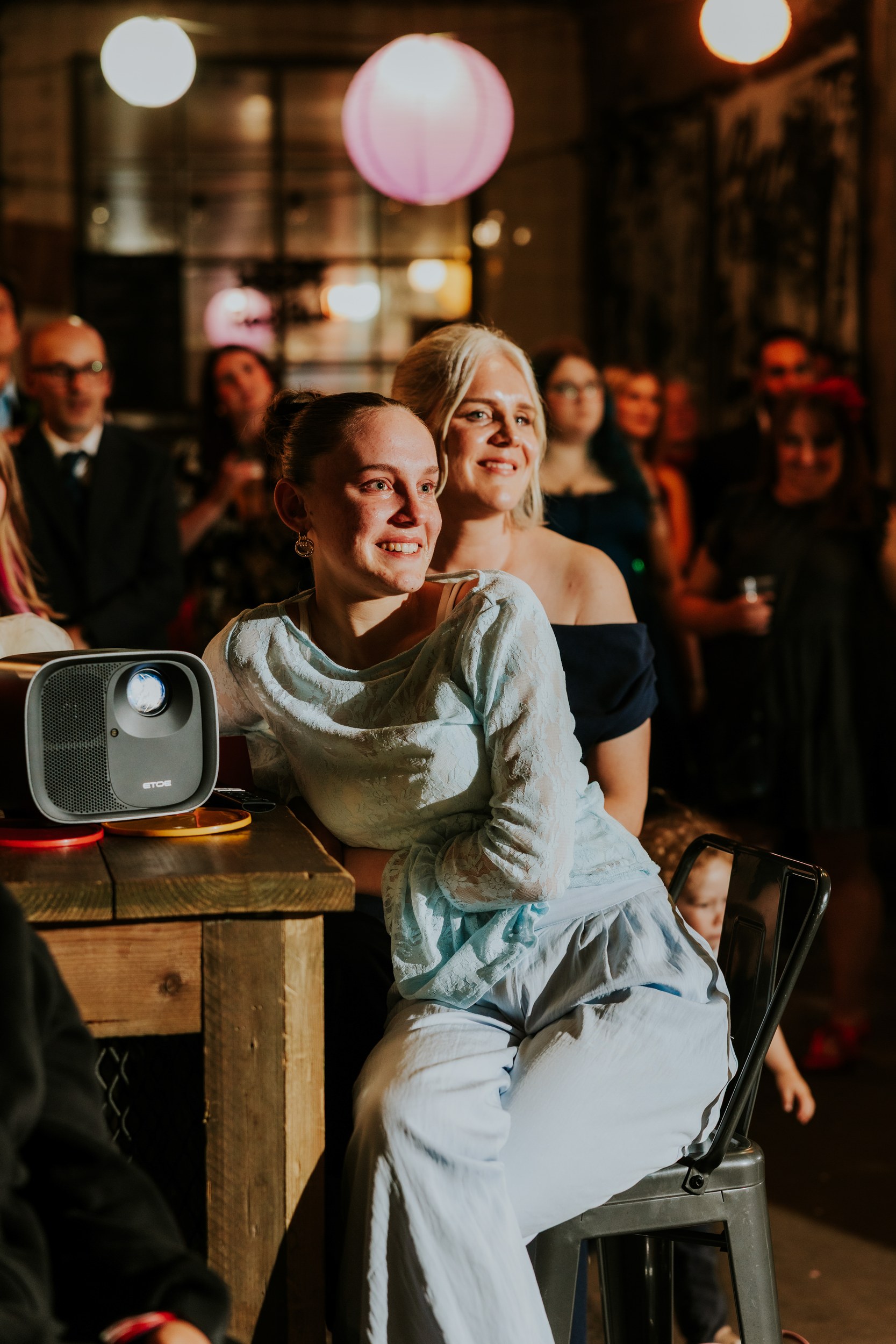 Daniel & Natalie’s Wedding at The Biscuit Factory 71 Two women smiling and watching a wedding presentation with a projector on a table and guests blurred behind.
