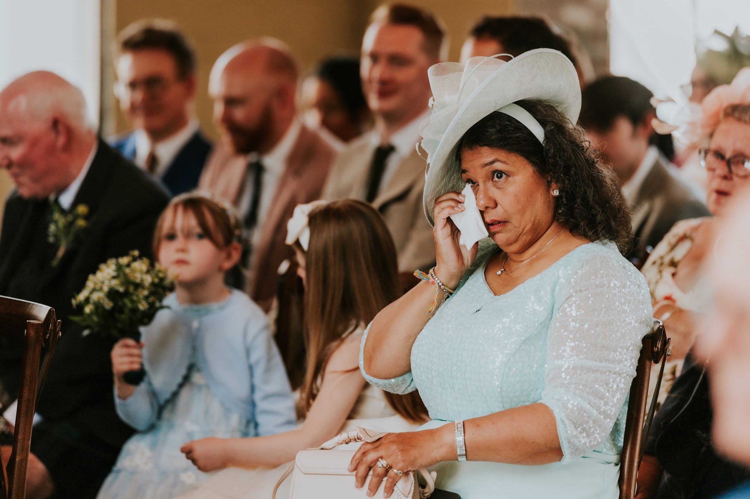Daniel & Natalie’s Wedding at The Biscuit Factory 21 Wedding guest in pale blue dress and hat dabbing tears with a tissue, children with bouquets and guests seated behind.