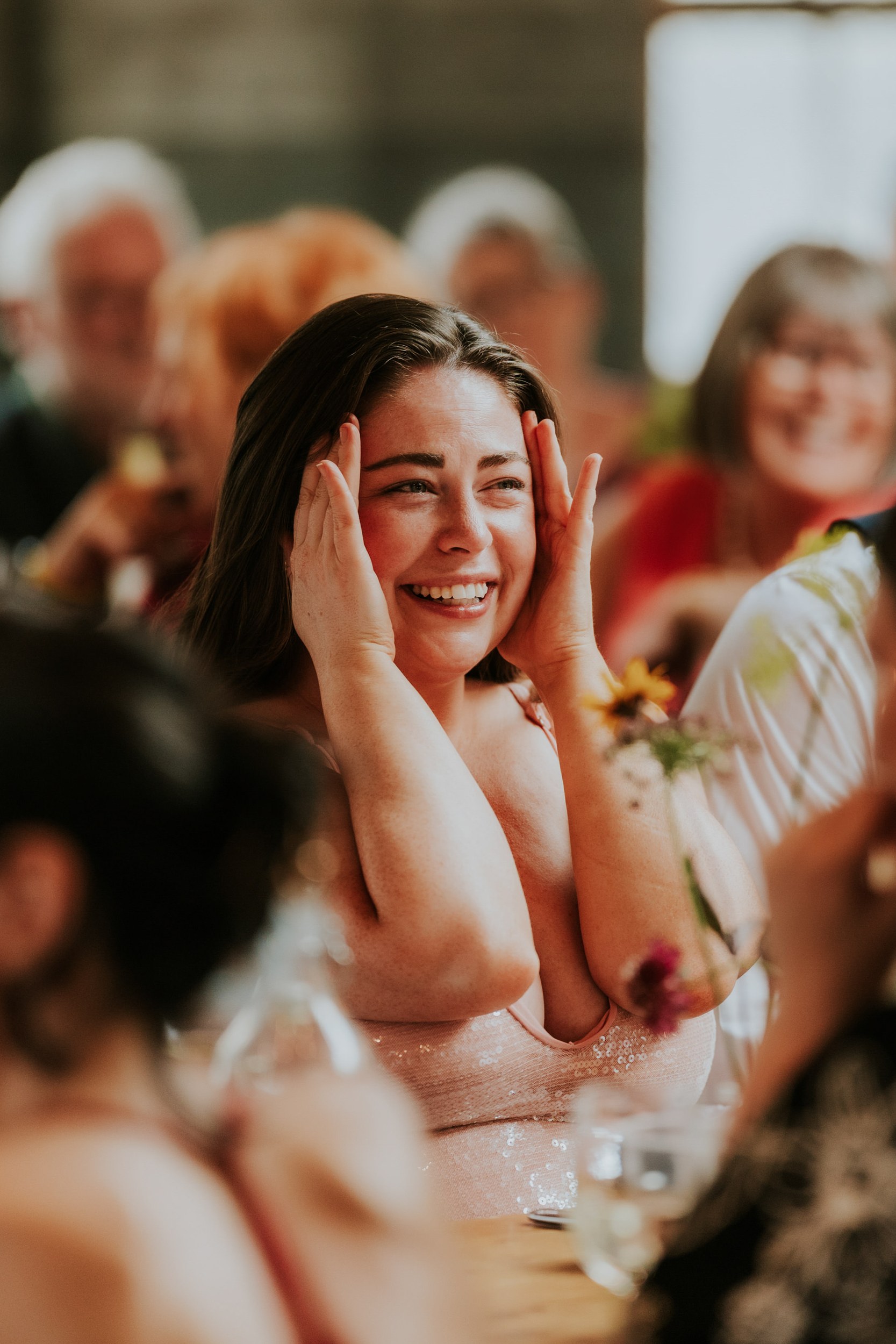 Daniel & Natalie’s Wedding at The Biscuit Factory 64 Wedding guest smiling with hands to her face at a reception table.
