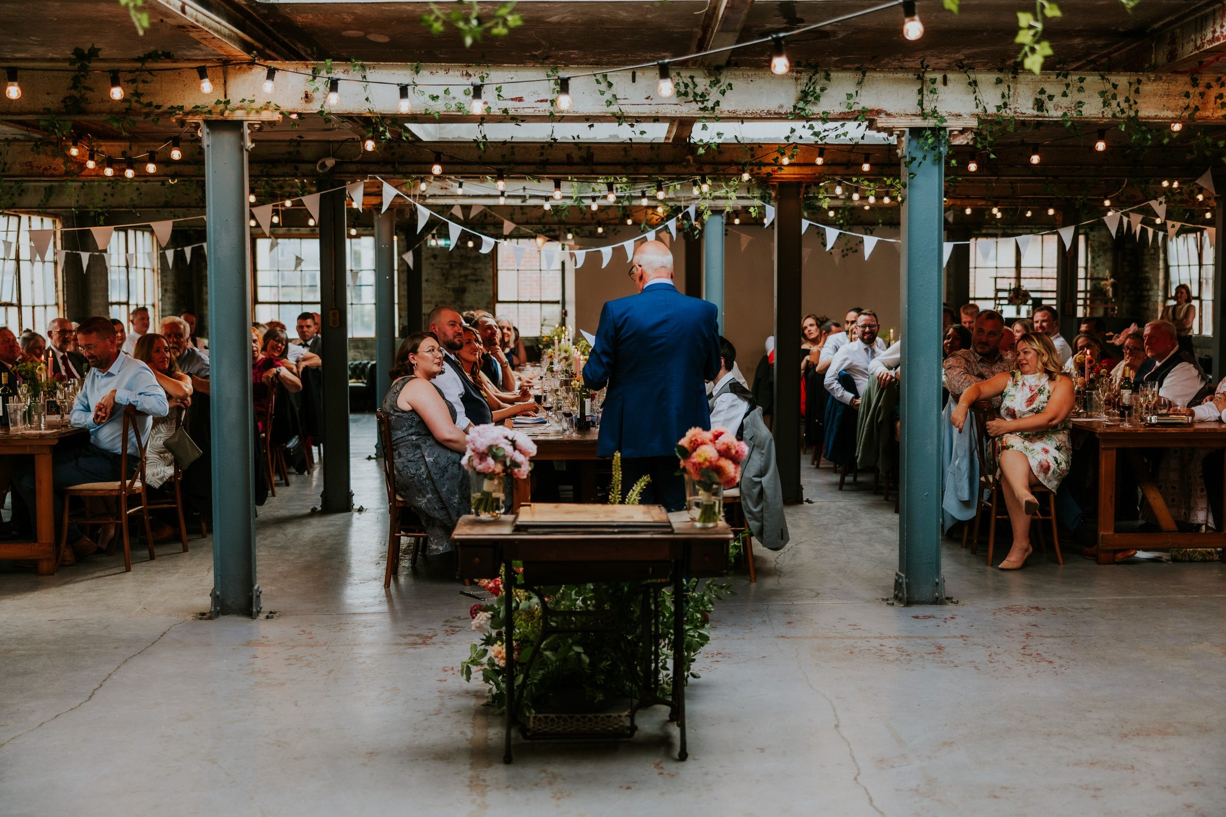 Daniel & Natalie’s Wedding at The Biscuit Factory 63 Wedding guests at The Biscuit Factory listening to a speaker under bunting and hanging lights.