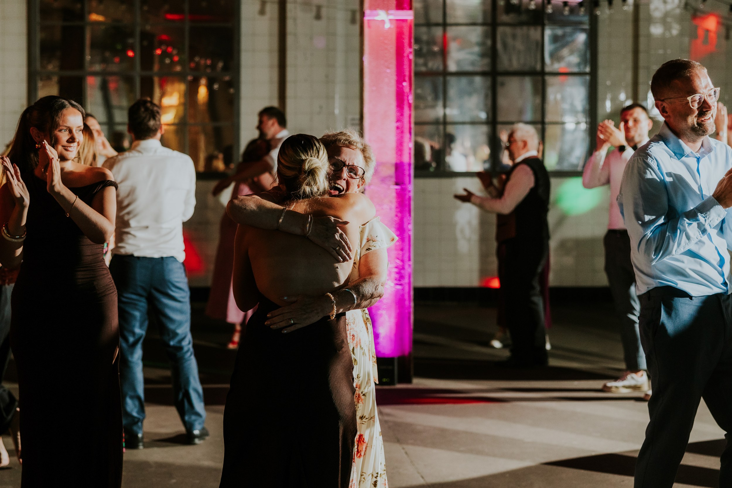 Daniel & Natalie’s Wedding at The Biscuit Factory 80 Wedding guests hugging and dancing by a pink-lit column on a dim reception dancefloor with large industrial windows behind.