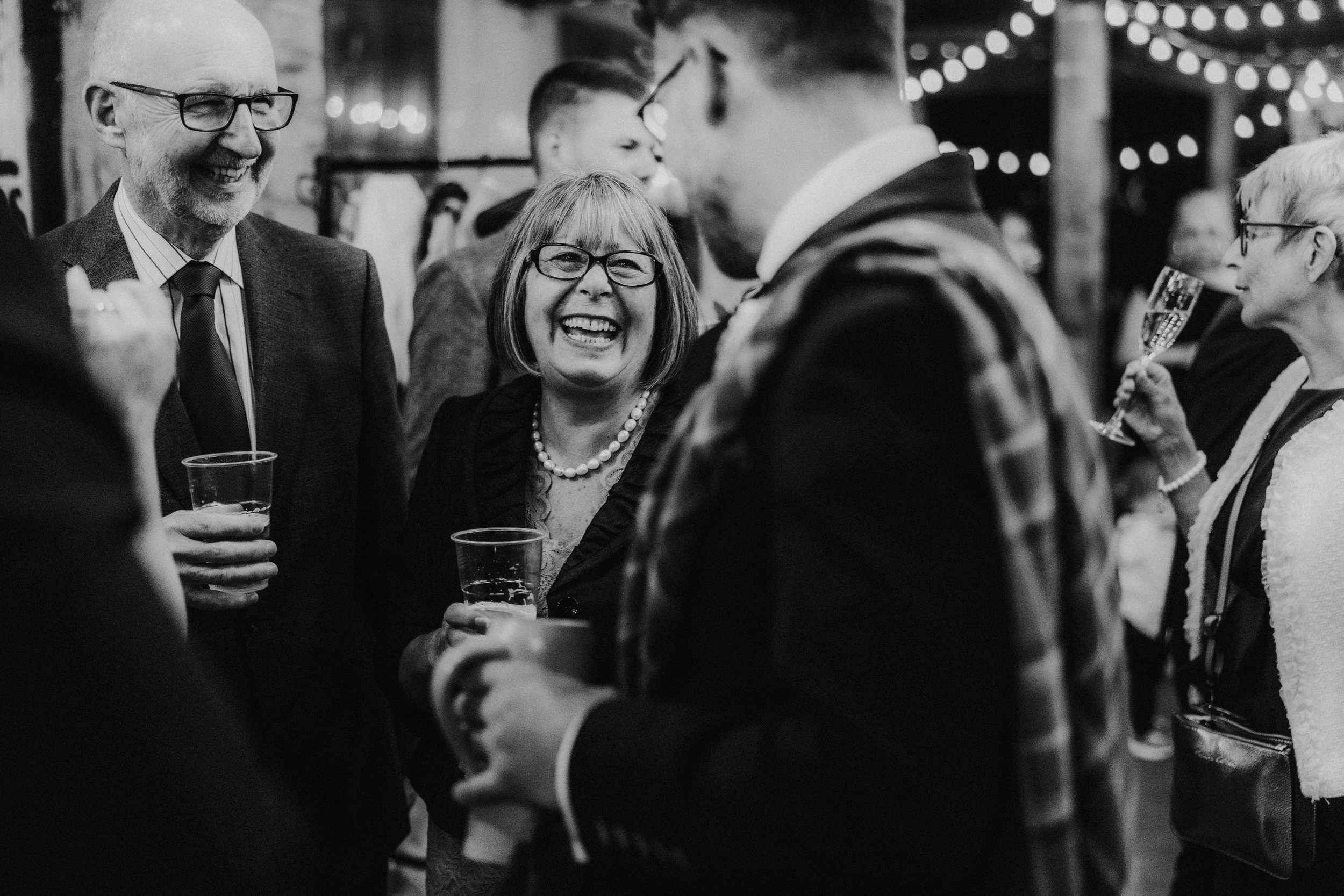Daniel & Natalie’s Wedding at The Biscuit Factory 38 Wedding guests laughing and holding drinks at an indoor reception with string lights in the background.