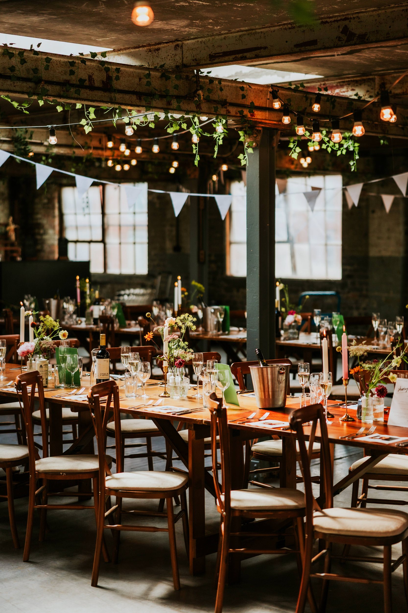 Daniel & Natalie’s Wedding at The Biscuit Factory 54 Wedding reception tables with candles, wine and flowers under bunting and hanging filament lights in an industrial room at The Biscuit Factory.
