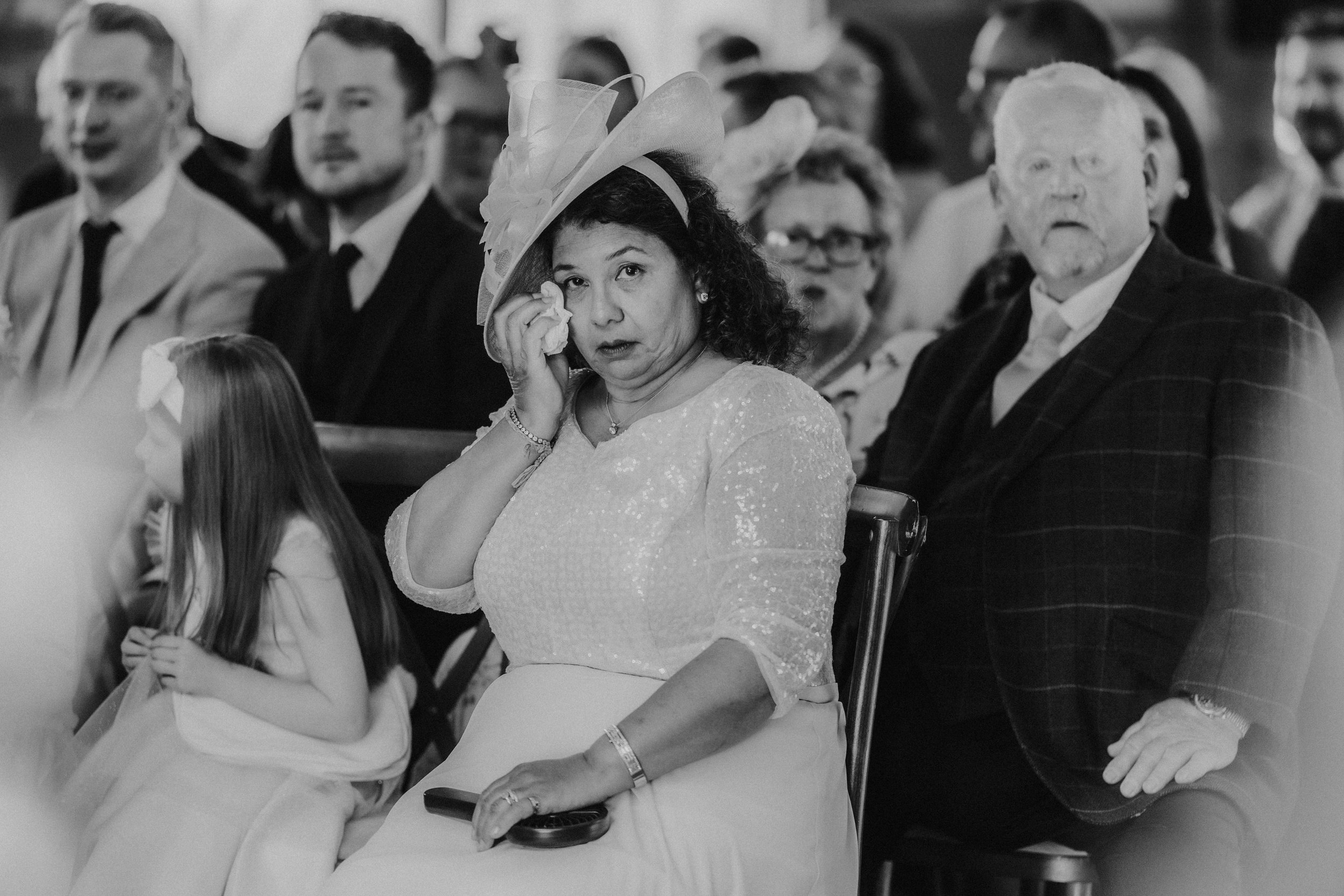 Daniel & Natalie’s Wedding at The Biscuit Factory 29 Woman in a formal hat wiping a tear with a tissue while seated among wedding guests at a ceremony.