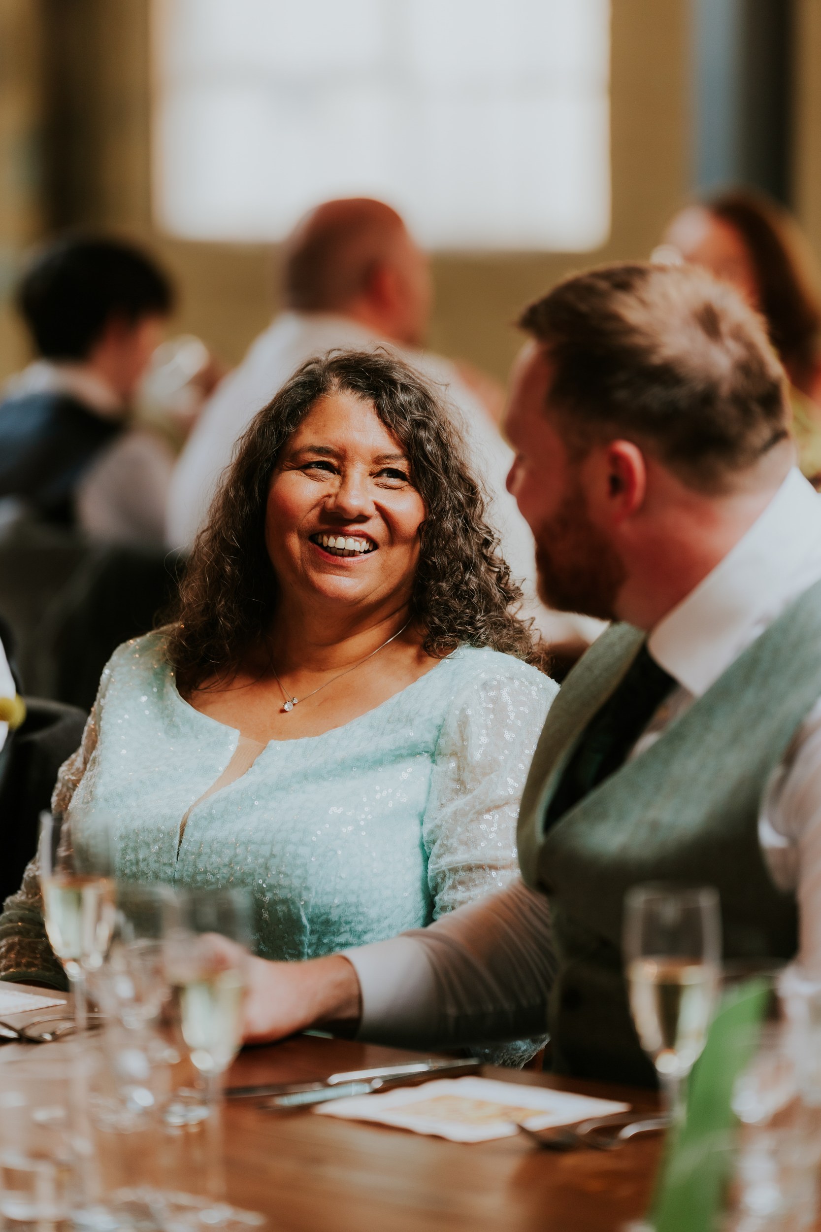 Daniel & Natalie’s Wedding at The Biscuit Factory 58 Woman in a pale blue sequinned dress smiling at a man across a wedding reception table.