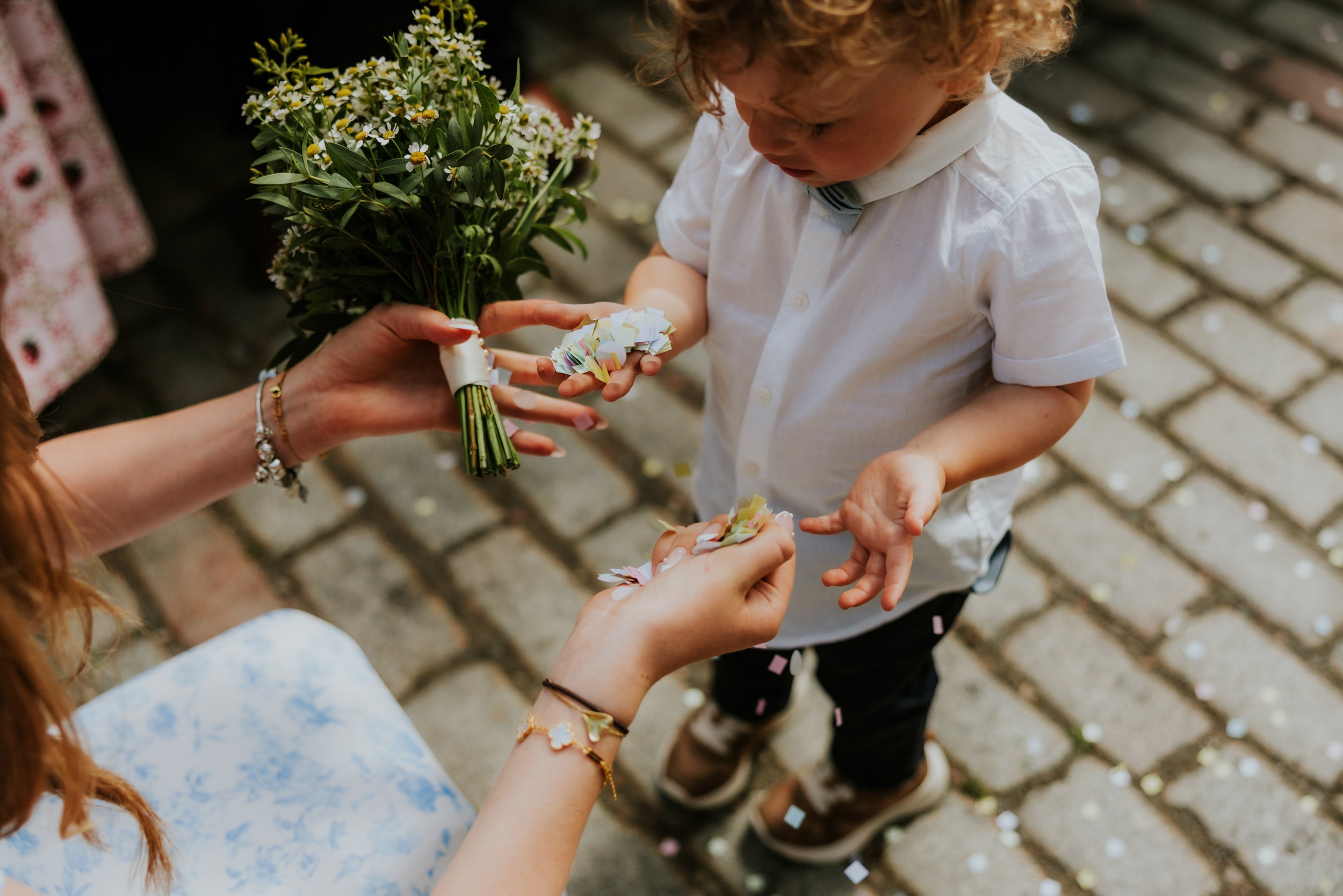 Daniel & Natalie’s Wedding at The Biscuit Factory 33 Young boy in white shirt receiving paper confetti from hands holding a small daisy bouquet on cobbles.