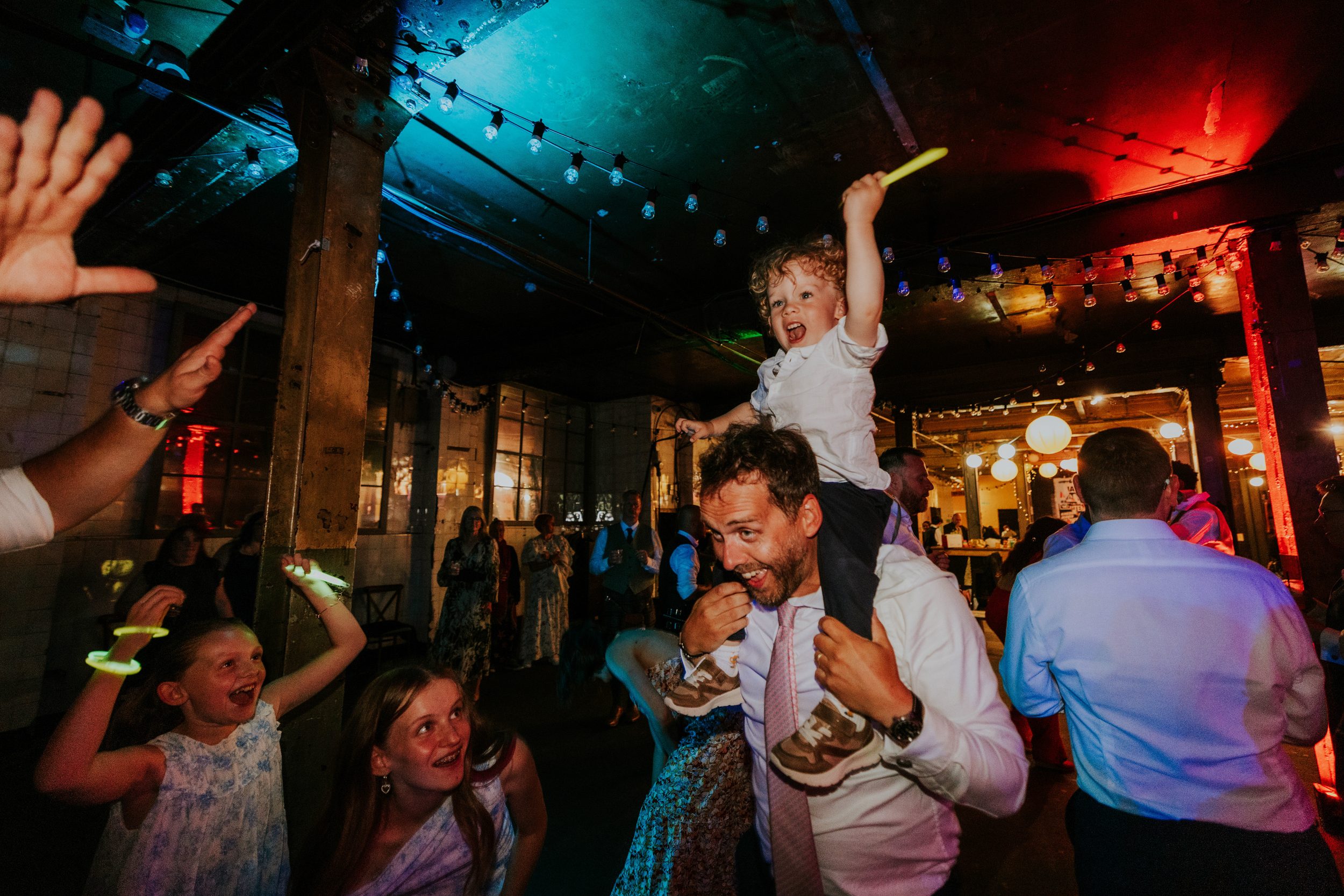 Daniel & Natalie’s Wedding at The Biscuit Factory 86 Young boy on a man's shoulders cheering with a glow stick on a crowded wedding dancefloor under string lights.