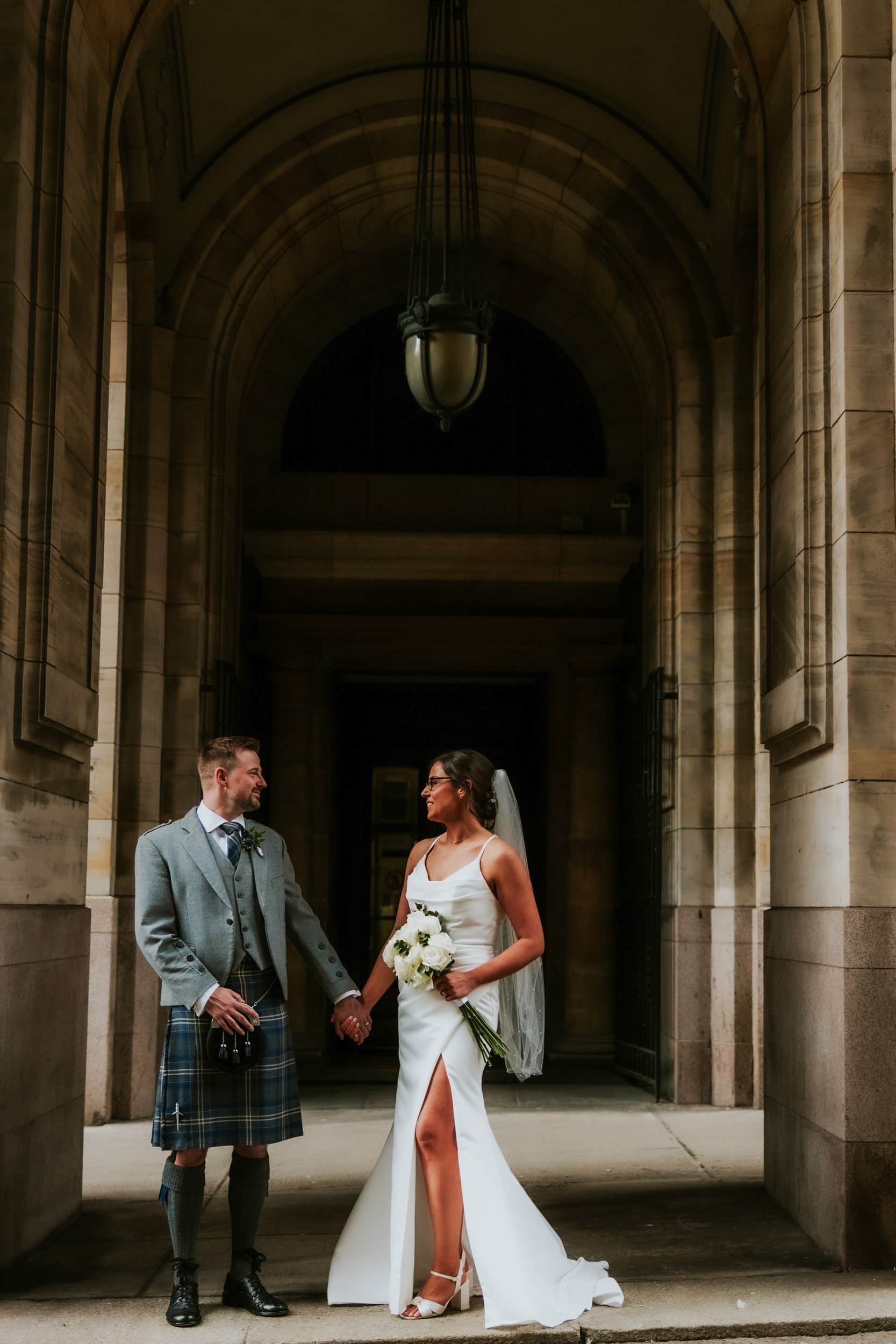 Alana & Kyle’s Glasgow Wedding — City Centre to The Bothy 17 Alana and Kyle holding hands under a stone archway; bride in white dress with bouquet, groom in grey jacket and kilt.