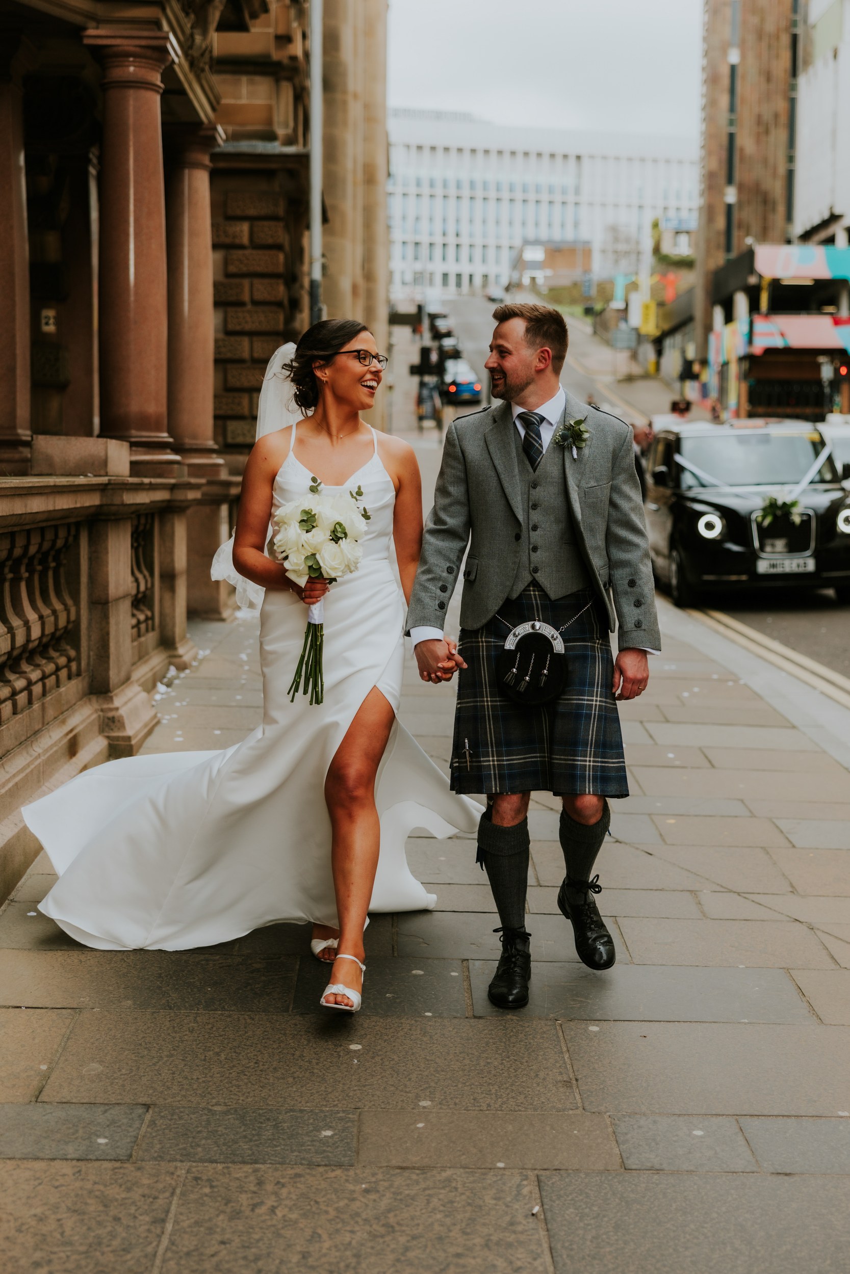 Alana & Kyle’s Glasgow Wedding — City Centre to The Bothy 12 Alana and Kyle walking on a Glasgow street; bride in white dress with bouquet, groom in kilt and grey jacket.