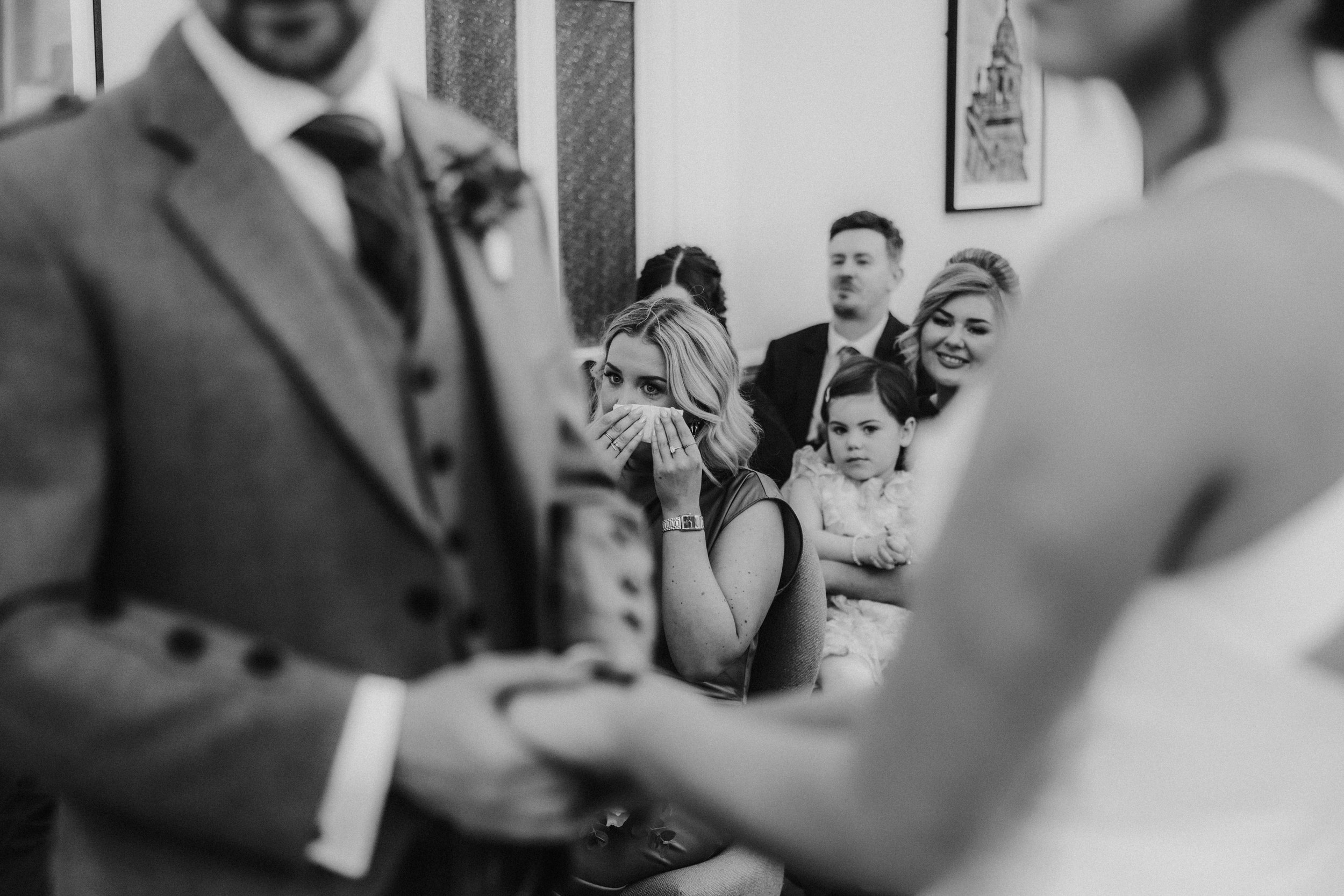 Alana & Kyle’s Glasgow Wedding — City Centre to The Bothy 7 Black-and-white ceremony photo: woman dabbing tears with tissue, child and guests watching, couple blurred in foreground.