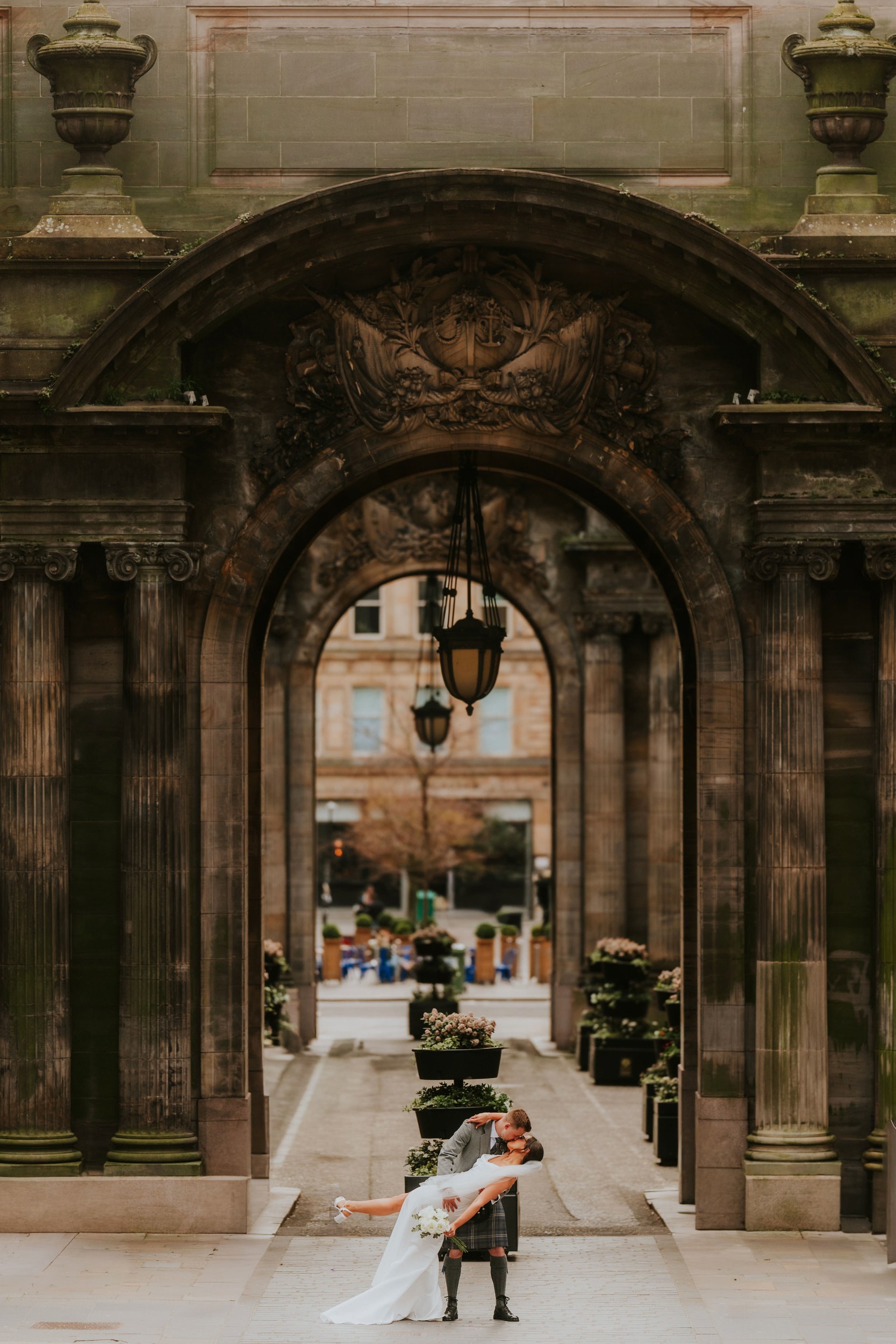 Alana & Kyle’s Glasgow Wedding — City Centre to The Bothy 14 Bride and groom kiss beneath stone archways at Glasgow City Chambers; groom in kilt, bride in white dress holding a bouquet.