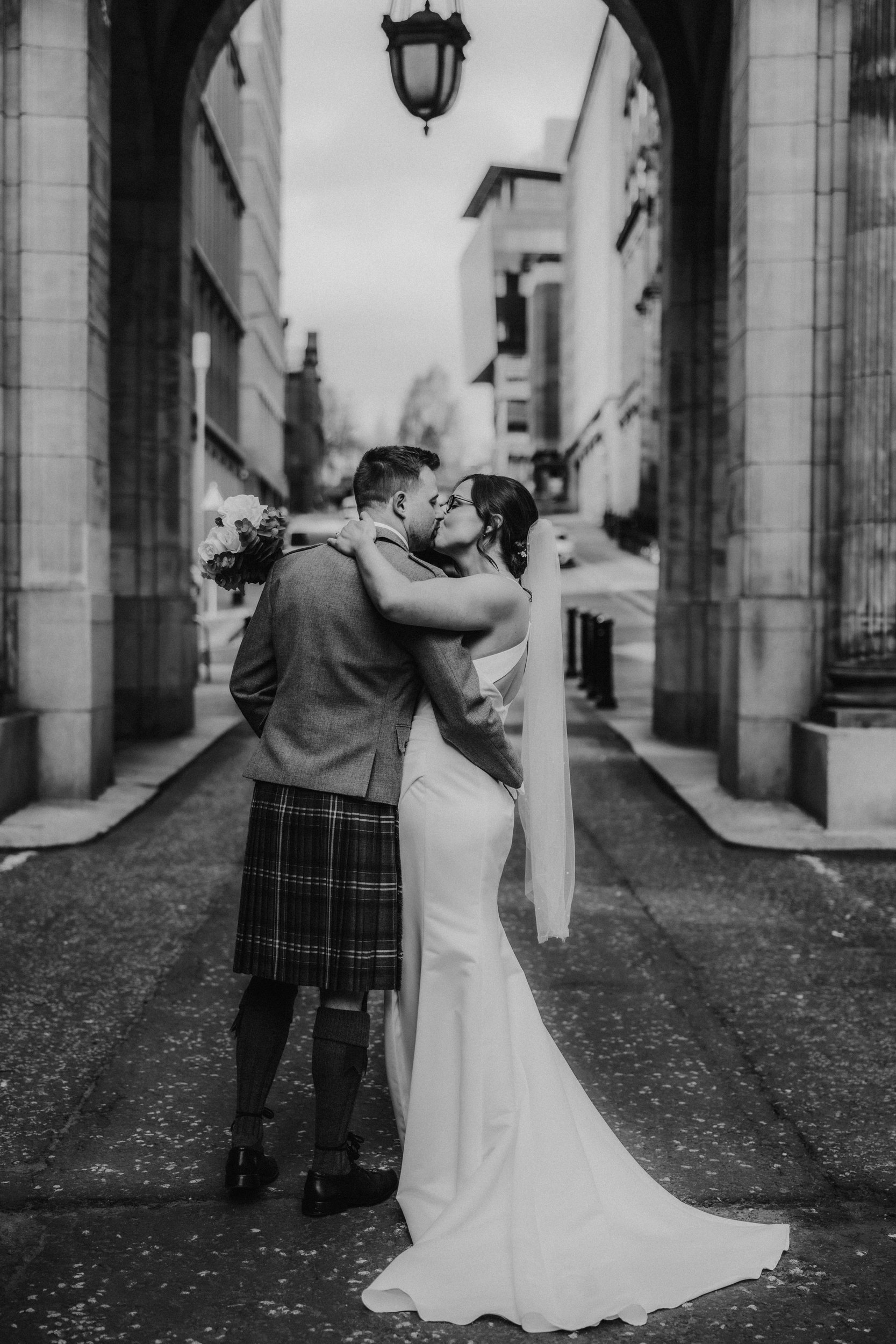 Alana & Kyle’s Glasgow Wedding — City Centre to The Bothy 16 Bride and groom kiss under a city stone arch; groom in kilt, bride in dress and veil with bouquet.