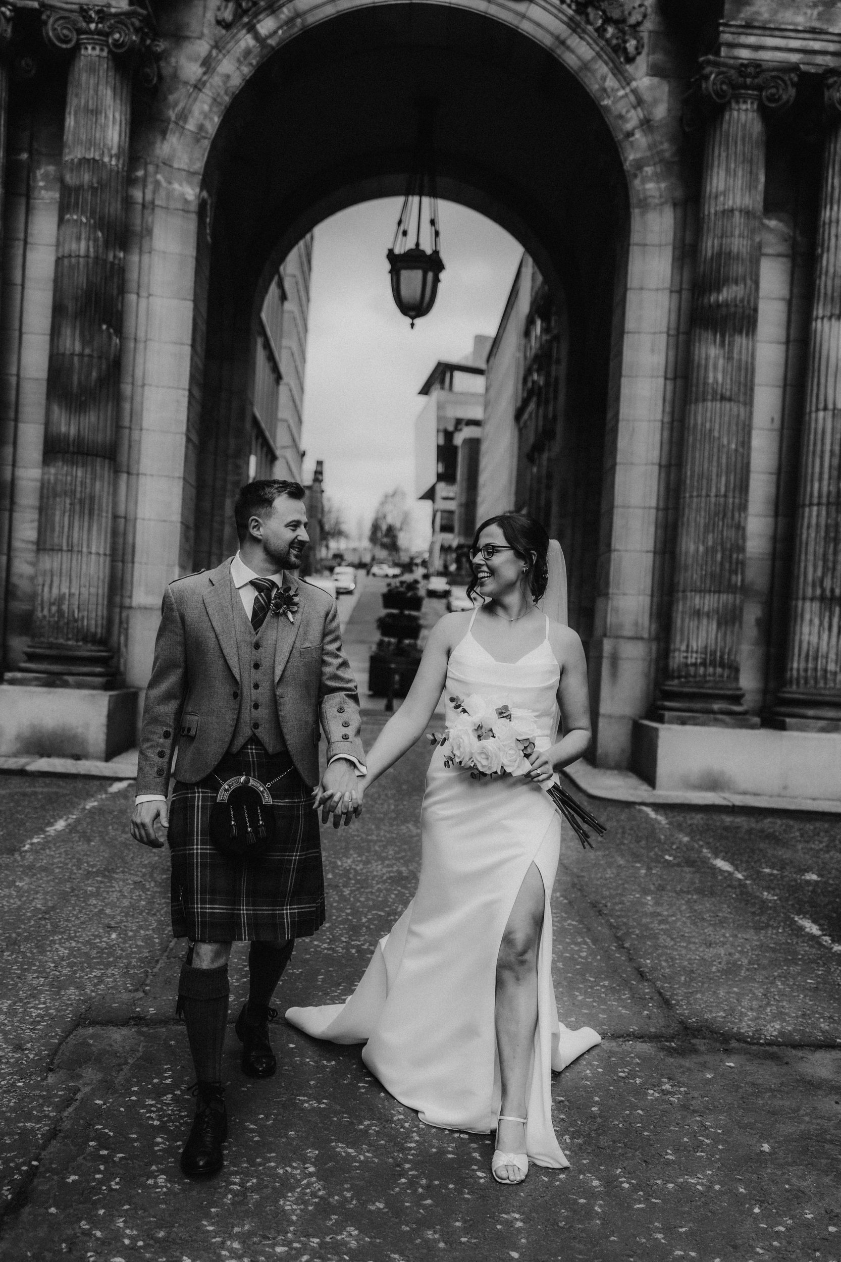 Alana & Kyle’s Glasgow Wedding — City Centre to The Bothy 18 Bride in sleeveless dress with bouquet and groom in kilt holding hands beneath a stone arch and columns in Glasgow city centre.